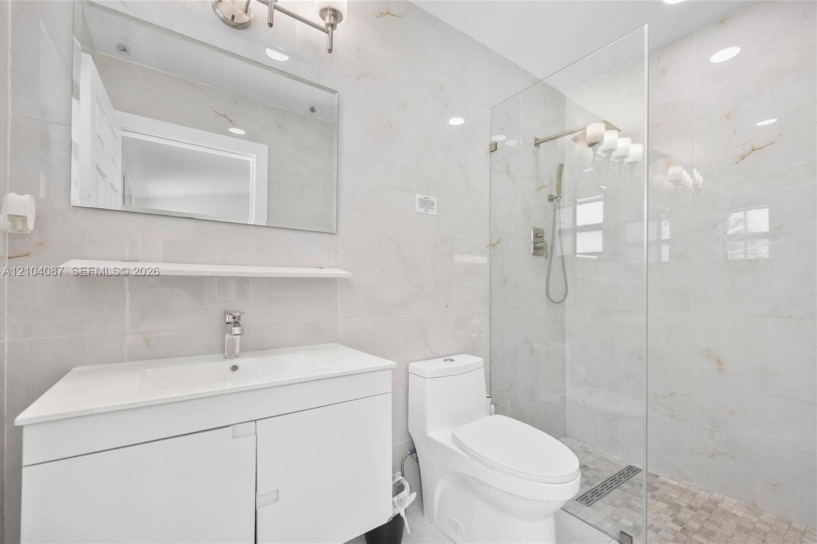 Bathroom featuring a glass-enclosed shower, wall-mounted shower system, white vanity with integrated sink, and white large-format wall tiles
