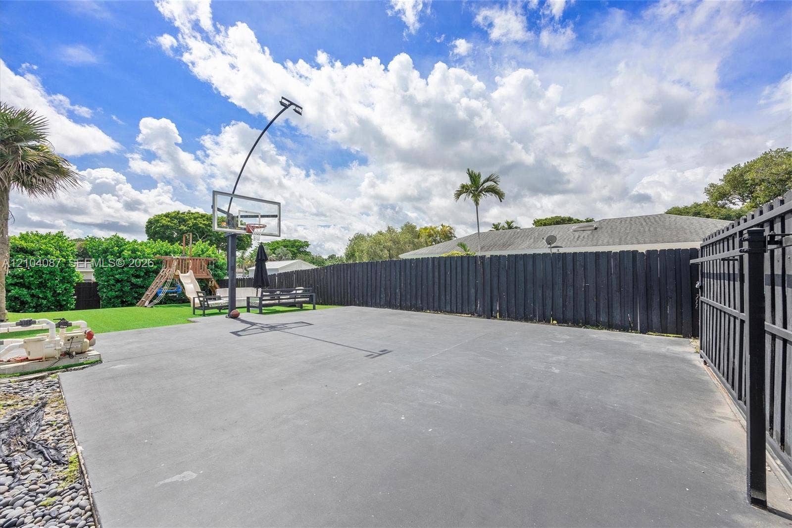 Expansive paved patio area featuring a basketball hoop, alongside a dark privacy fence and a gated entry