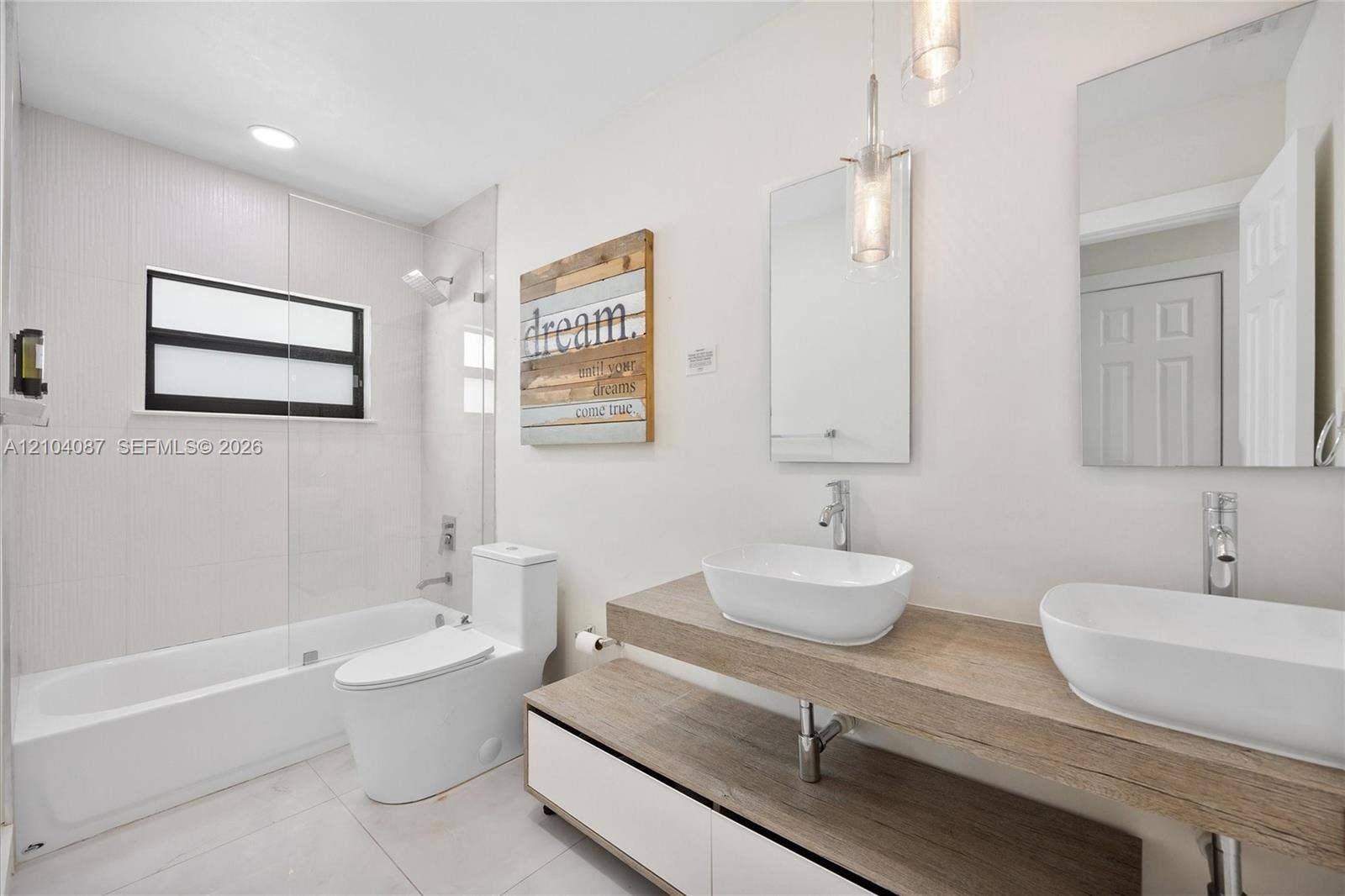 8840 SW 181st Terrace, Palmetto Bay, FL 33157 Dual vessel sinks with chrome faucets, wood-finish vanity with integrated storage, full-sized bathtub with glass enclosure, white tile flooring, and recessed lighting