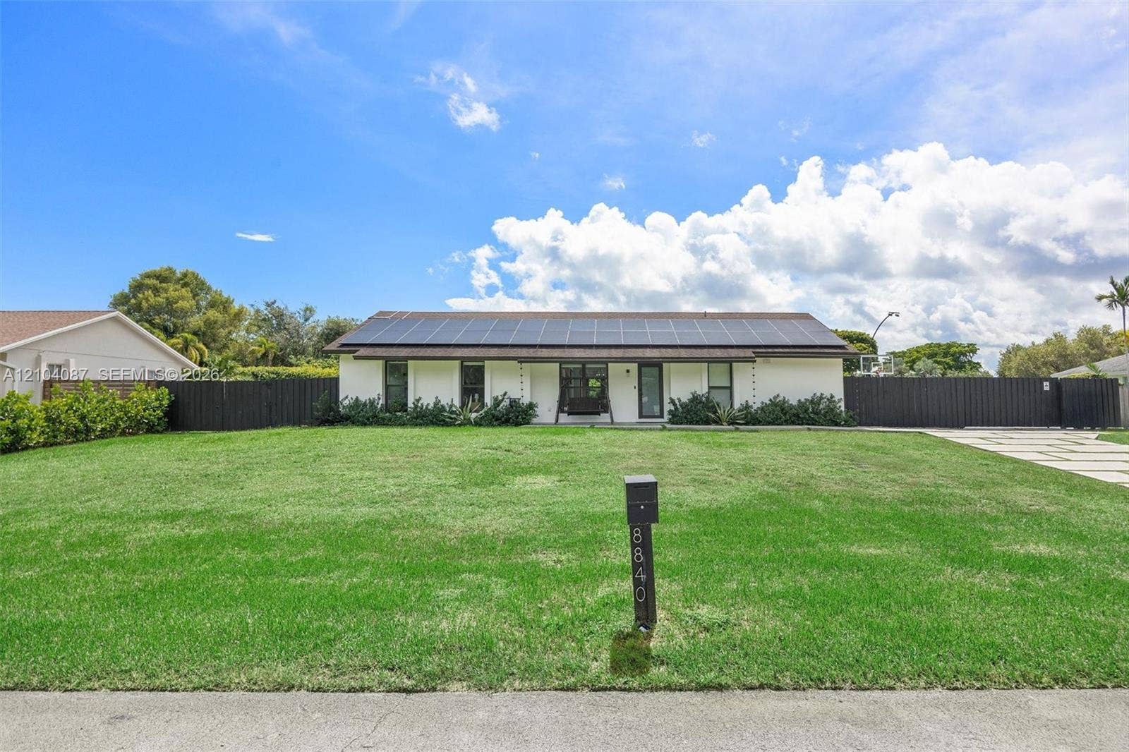 8840 SW 181st Terrace, Palmetto Bay, FL 33157 White exterior with dark trim accents, solar panel array, black fence, and a paver driveway
