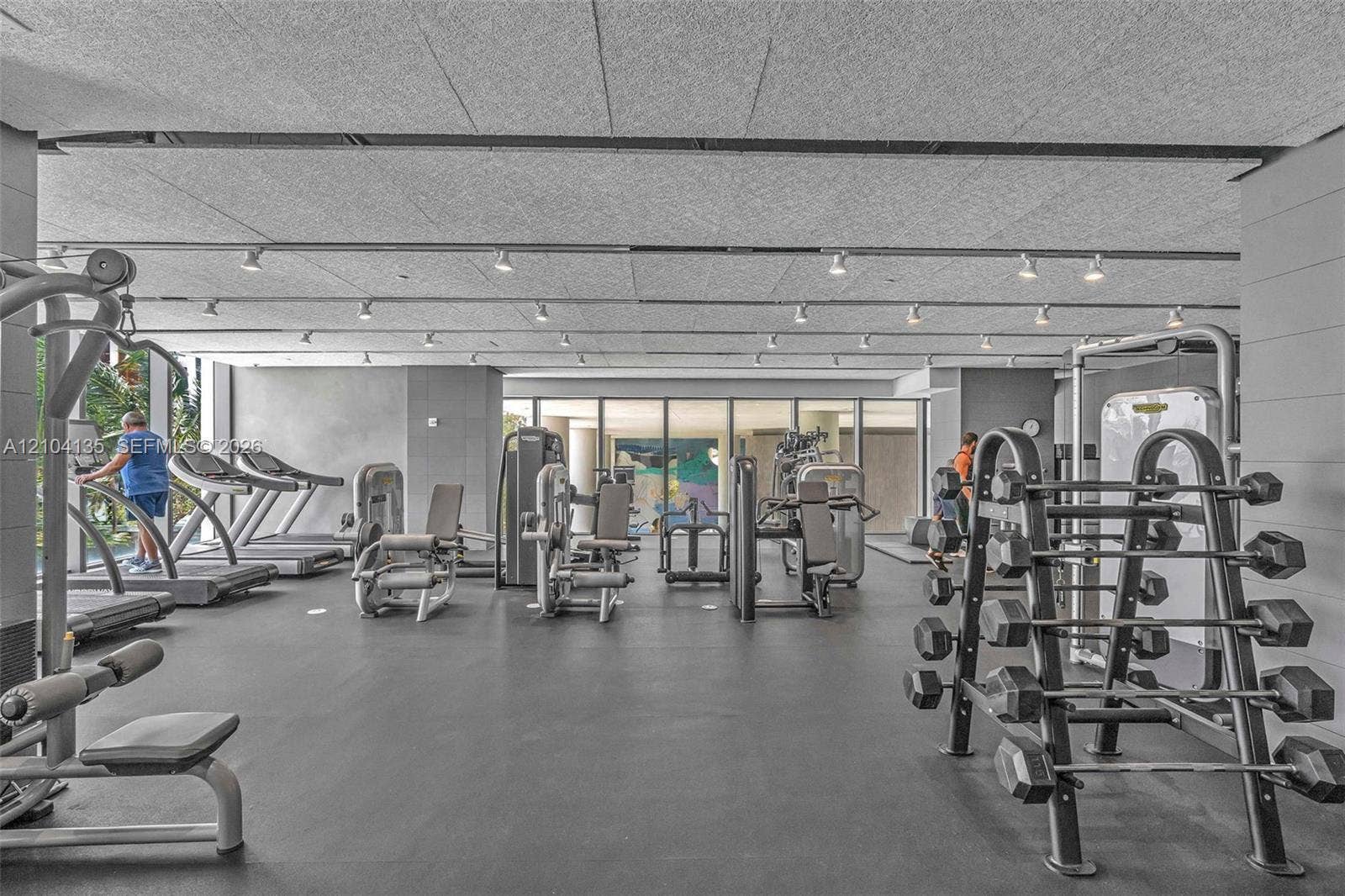 Fitness center featuring industrial-style ceiling tiles, expansive floor-to-ceiling windows, recessed track lighting, and durable rubberized flooring