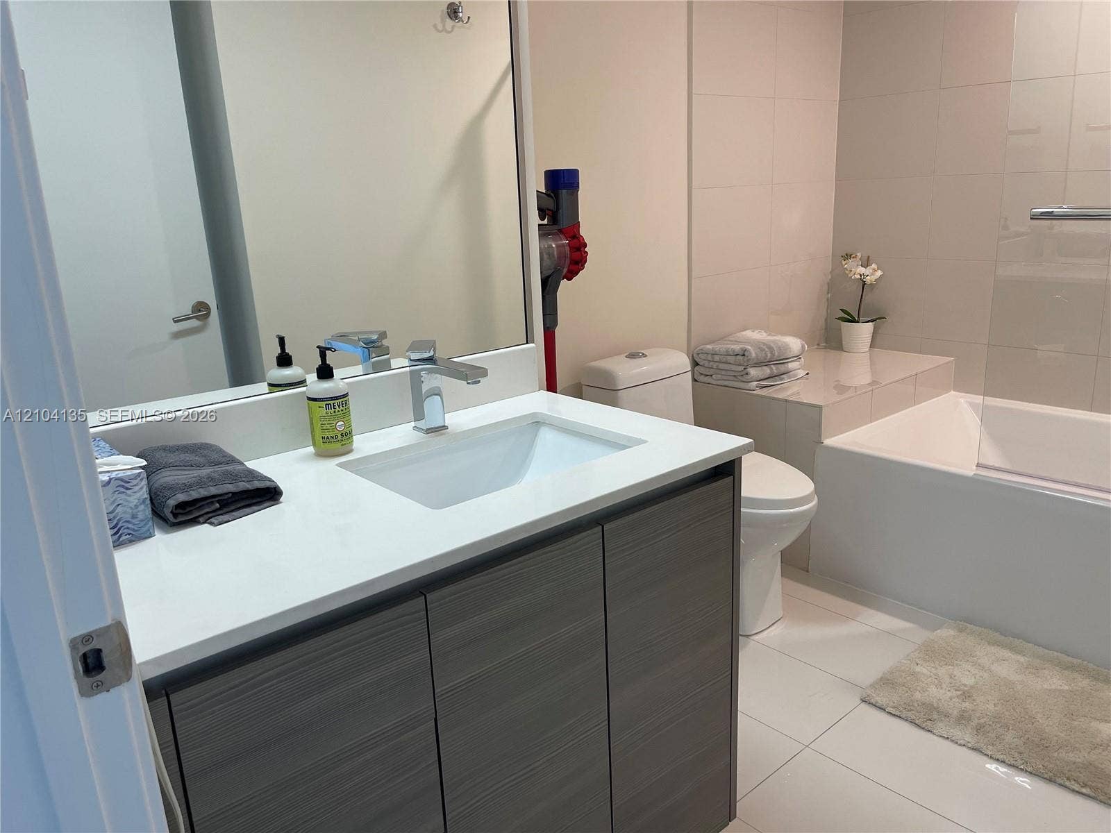 Bathroom featuring a vanity with wood-finish cabinetry, white countertop with integrated sink, chrome faucet, and a large-format tiled bathtub enclosure with a glass shower screen