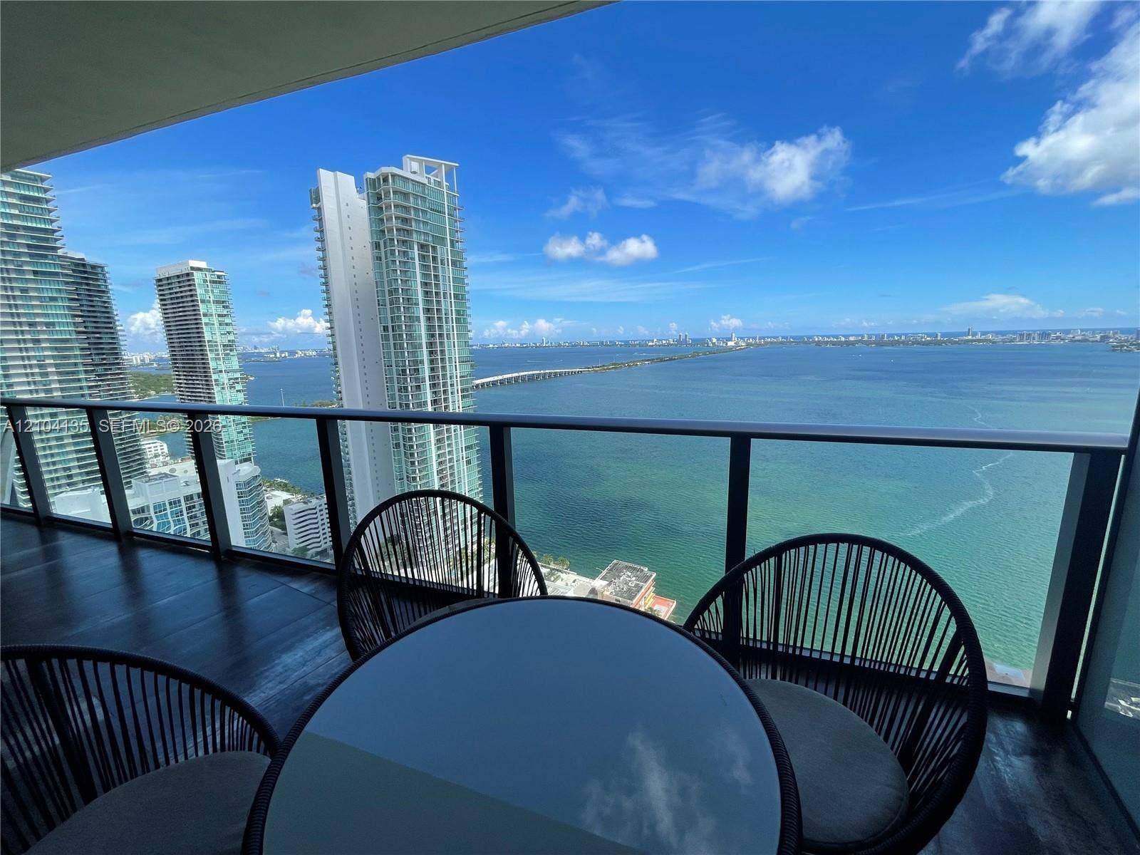 Covered balcony featuring dark-toned flooring and a black metal railing with glass panels, overlooking the water and surrounding high-rise buildings