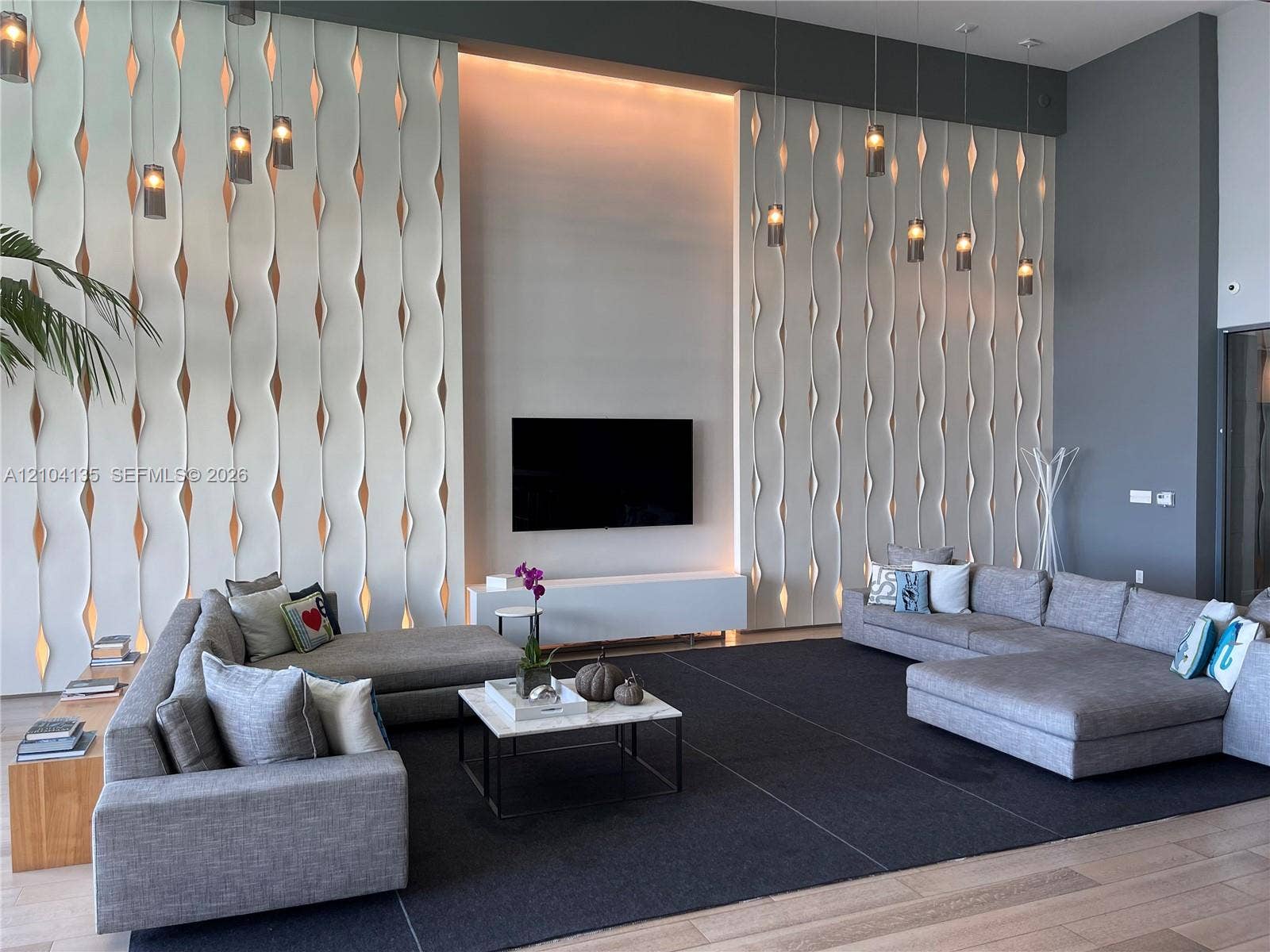 High-ceiling room featuring custom, floor-to-ceiling sculptural wall panels with integrated lighting, minimalist wall-mounted media console, and a series of hanging pendant light fixtures