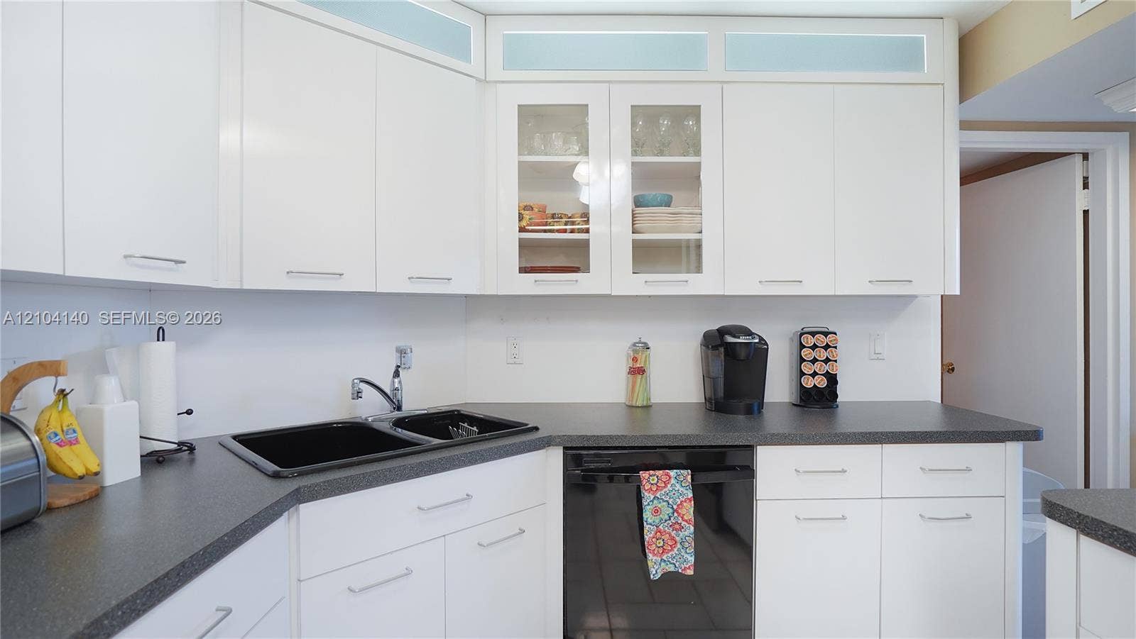 111 N Pompano Beach Boulevard, #304, Pompano Beach, FL 33062 Kitchen featuring white cabinetry with glass-front display panels, transom accent windows, dark-toned countertops, and a dual-basin sink with a chrome faucet