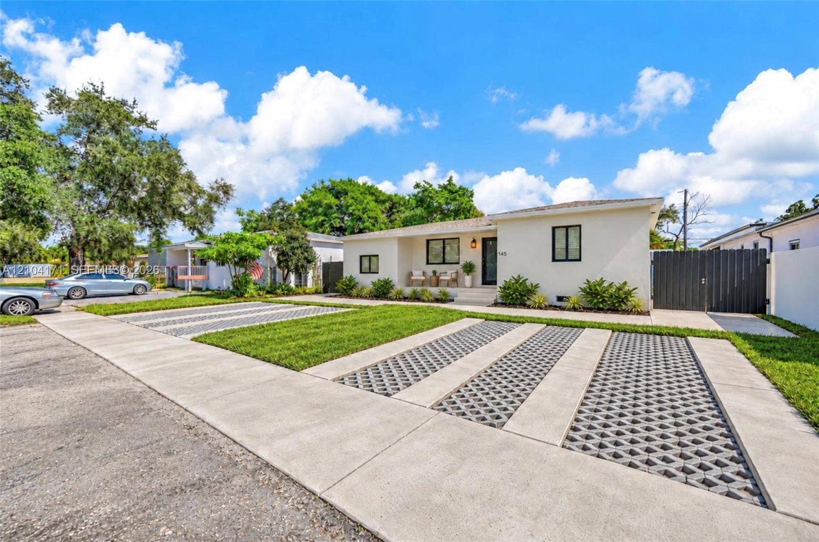 745 NW 32nd Avenue, Miami, FL 33125 Single-story residence featuring light-colored exterior siding, multi-vehicle parking area with geometric paver inserts, manicured front lawn, and dark-finished privacy fencing