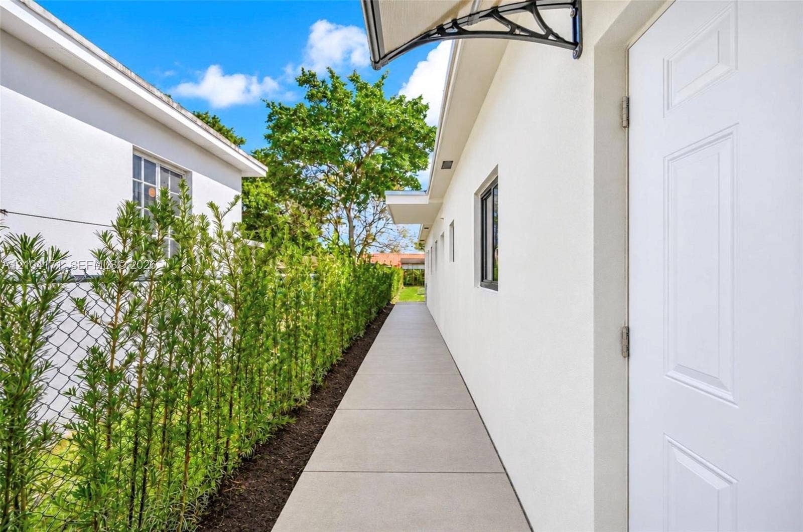 745 NW 32nd Avenue, Miami, FL 33125 Exterior walkway featuring concrete paving, smooth stucco siding, a metal door awning, and a tall privacy hedge