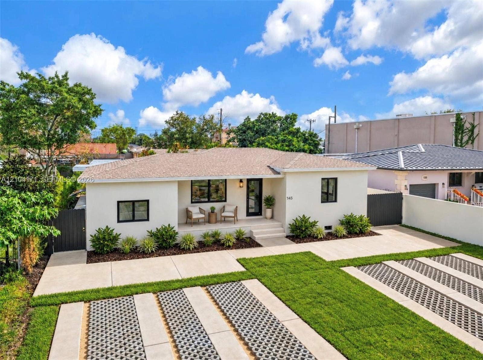 745 NW 32nd Avenue, Miami, FL 33125 Single-story residence featuring a crisp exterior finish, a covered entryway, a roof with shingle-style materials, dark-framed windows, and a multi-section concrete driveway with integrated turf grid pavers