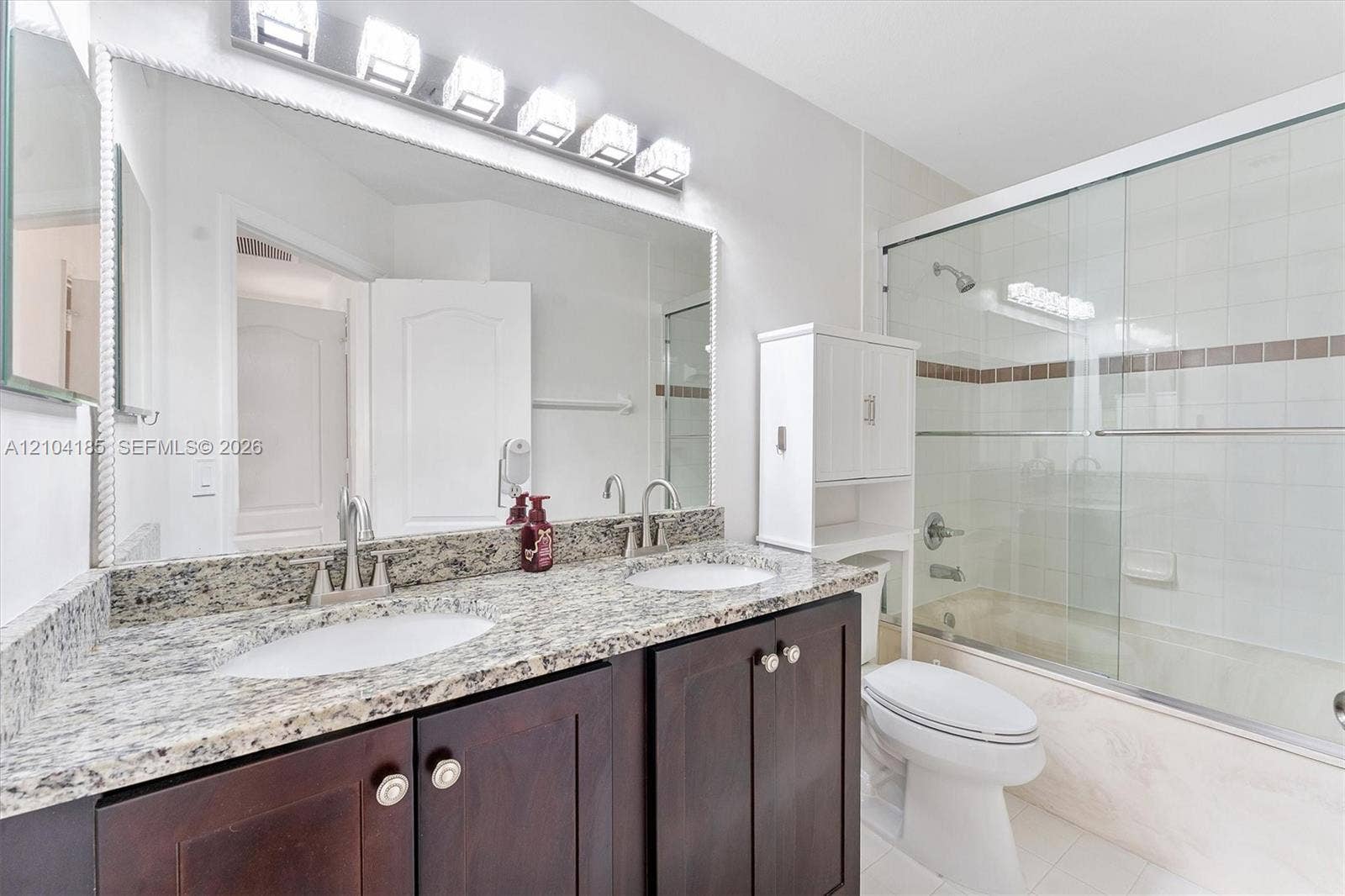 Bathroom featuring a dual-sink vanity with stone-finish countertops, dark wood-finish cabinetry, a full-width mirror, and a shower-tub combination with glass enclosure