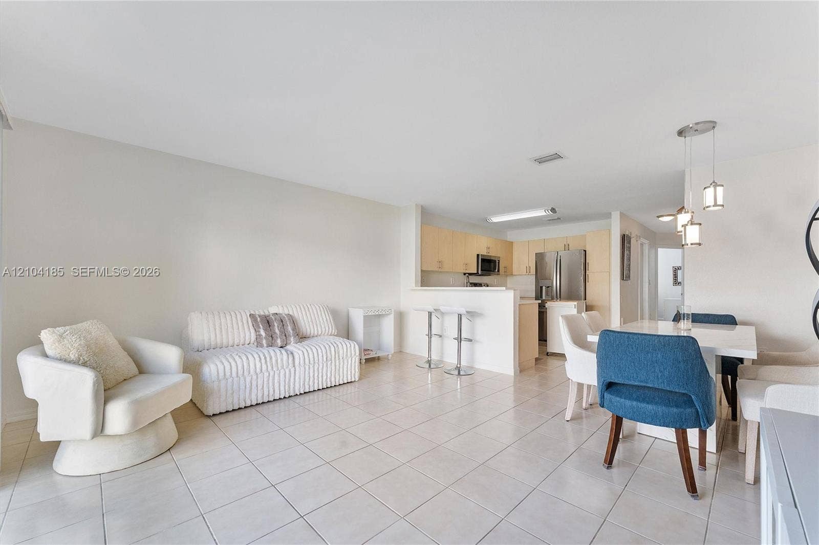 Open-concept interior featuring large-format ceramic tile flooring, neutral wall finishes, a kitchen peninsula with integrated breakfast bar seating, stainless steel appliances, and minimalist pendant lighting