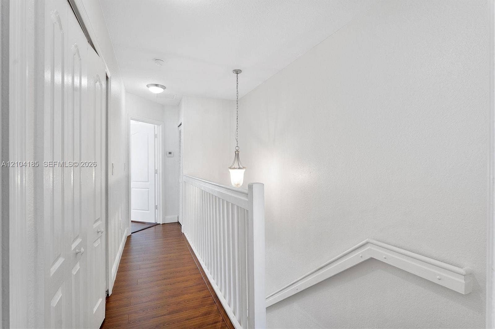 Hallway featuring wood-finish flooring, white paneled closet doors, a decorative pendant light fixture, and a white balustrade