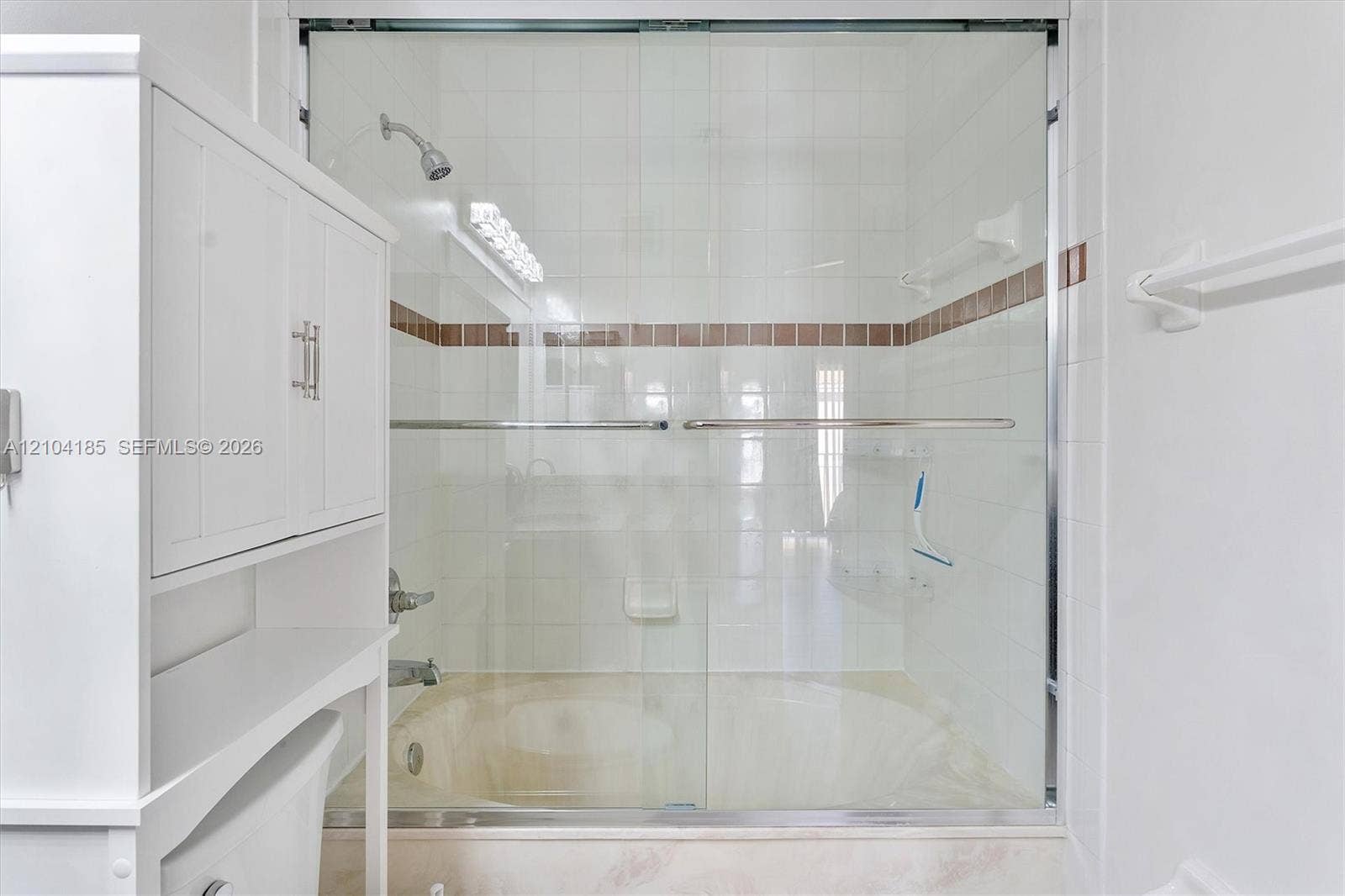 Shower and tub combination featuring a sliding glass enclosure, white ceramic tile surround with a decorative horizontal inlay, built-in shelving, and white cabinetry