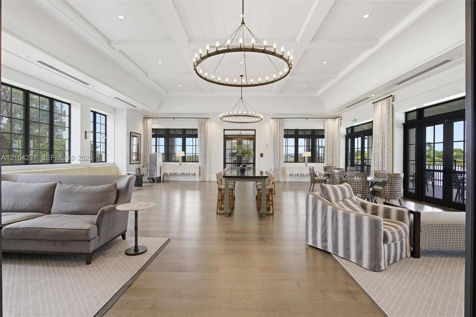 Expansive room featuring coffered ceilings, oversized circular chandeliers, wood-finish flooring, multi-pane windows with black frames, and glass-paned double doors