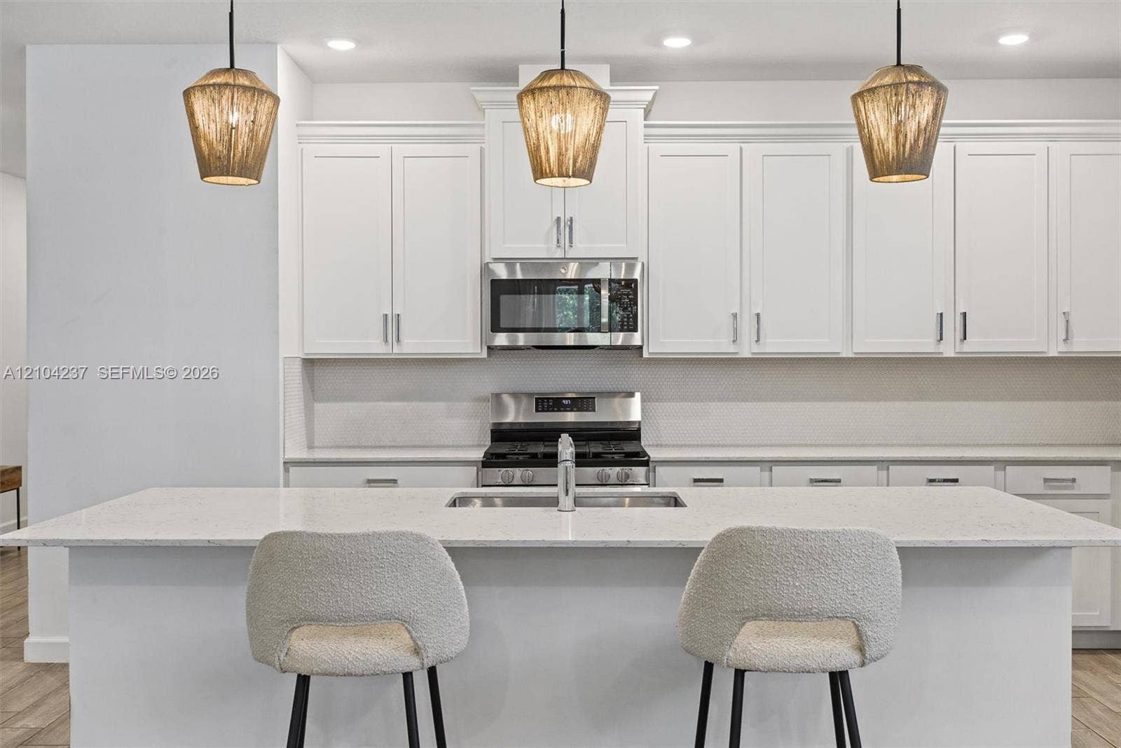 Kitchen featuring white Shaker-style cabinetry, a stone-composite countertop island with an integrated stainless steel sink, stainless steel built-in microwave, stainless steel gas range, and three hanging textured pendant lights