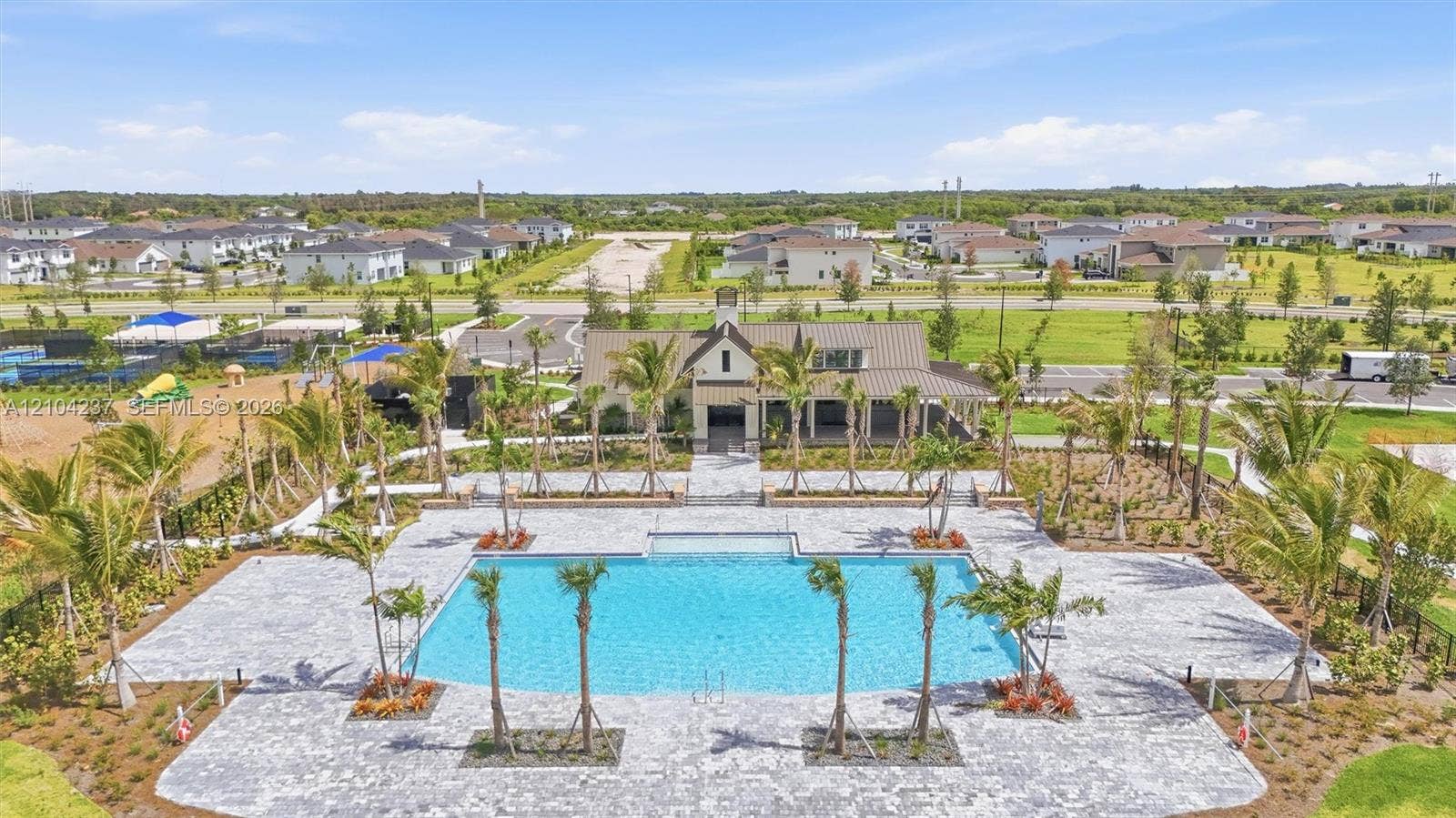 Community swimming pool featuring a paved deck, surrounding palm trees, adjacent clubhouse with a covered veranda, and multi-sport courts