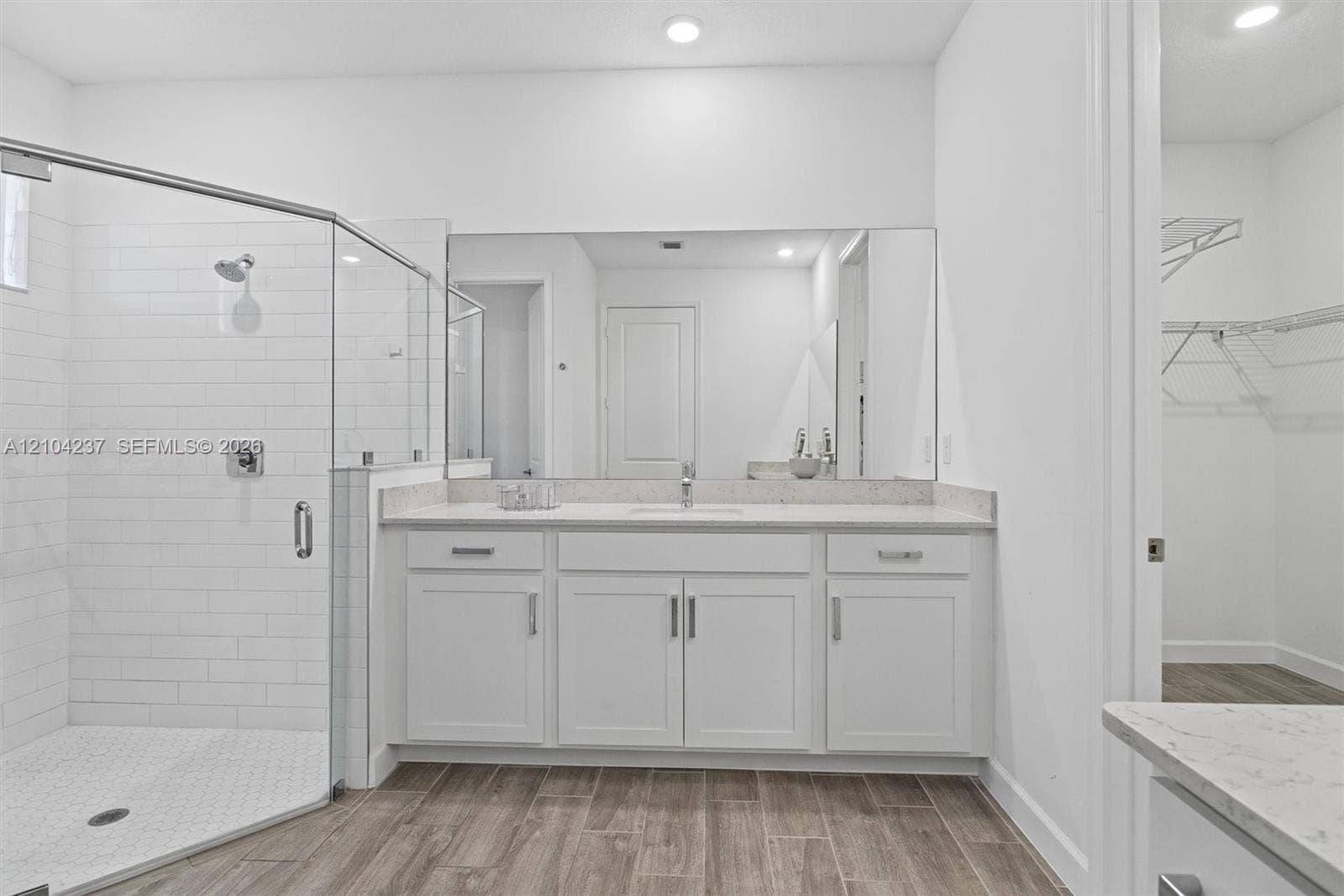 Bathroom featuring white cabinetry, light-toned stone-finish countertop with integrated sink, walk-in shower with glass enclosure and subway tile surround, full-width wall mirror, and wood-finish flooring