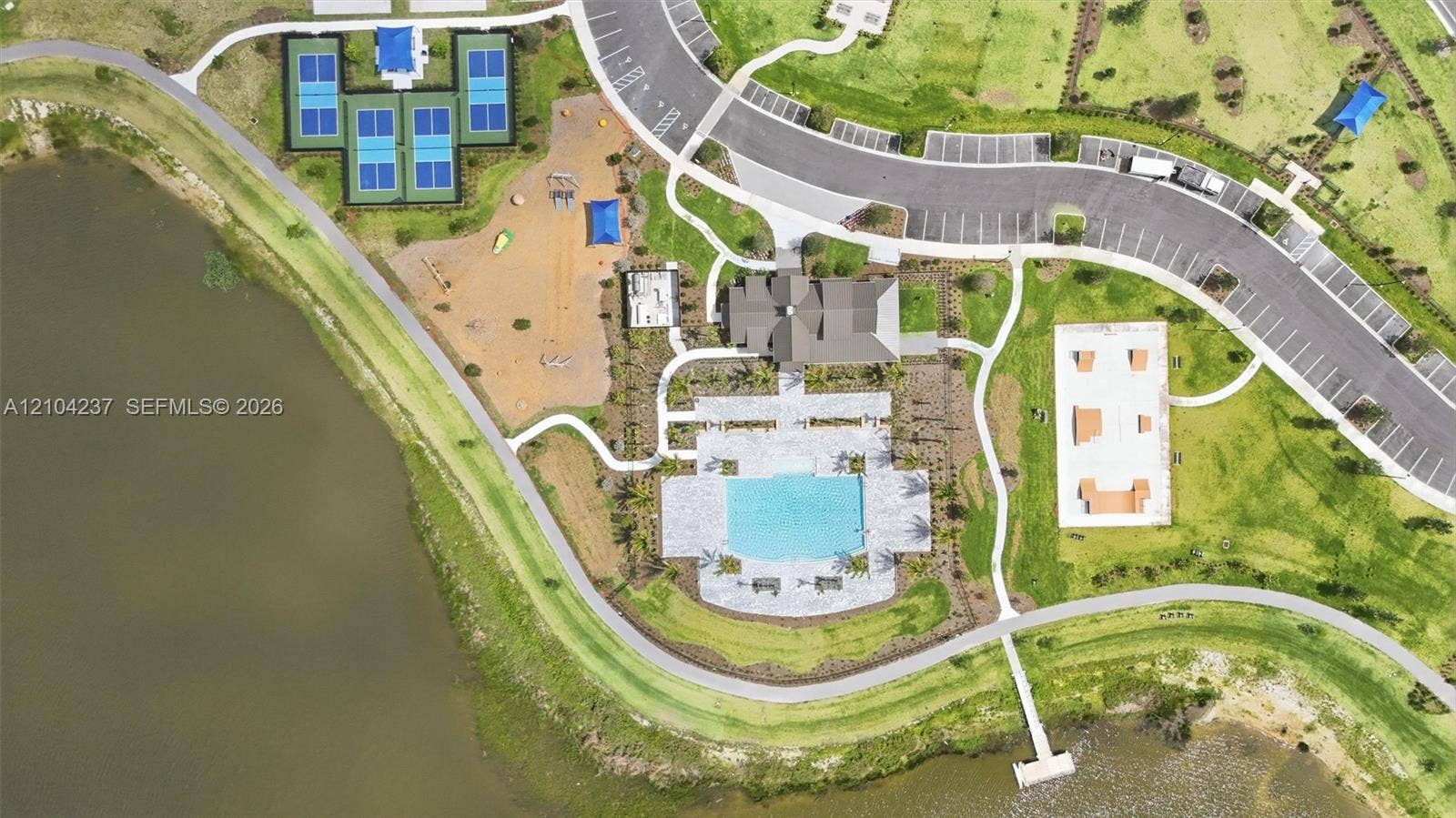 Aerial perspective of recreation area featuring a swimming pool with paved deck, four tennis courts, a playground, skate park, and shoreline access