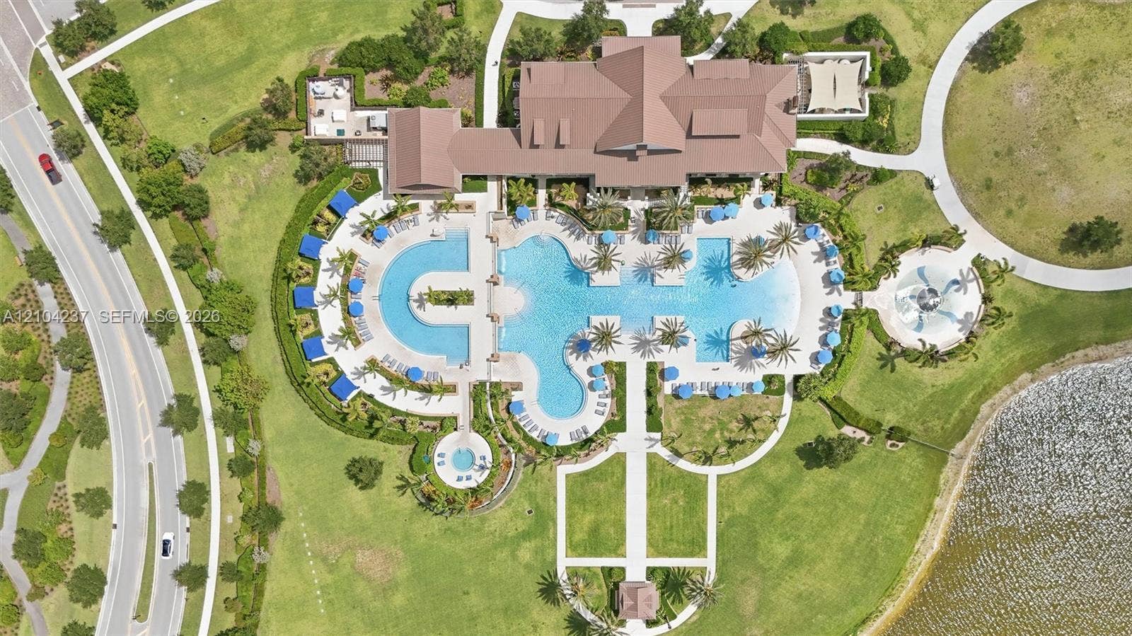 Aerial perspective of property amenities featuring a multi-section outdoor swimming pool, a circular spa, landscaped grounds with mature palm trees, concrete walking paths, and a dedicated pavilion