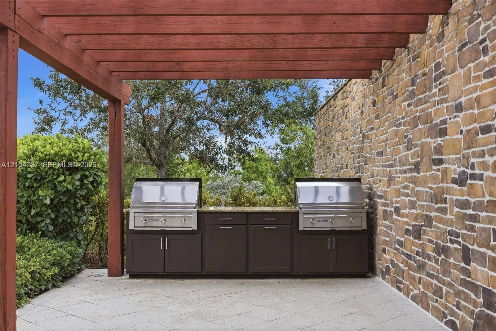 Outdoor kitchen featuring a stone-clad wall, natural wood pergola, dual stainless steel grills, dark cabinetry, and stone paver flooring