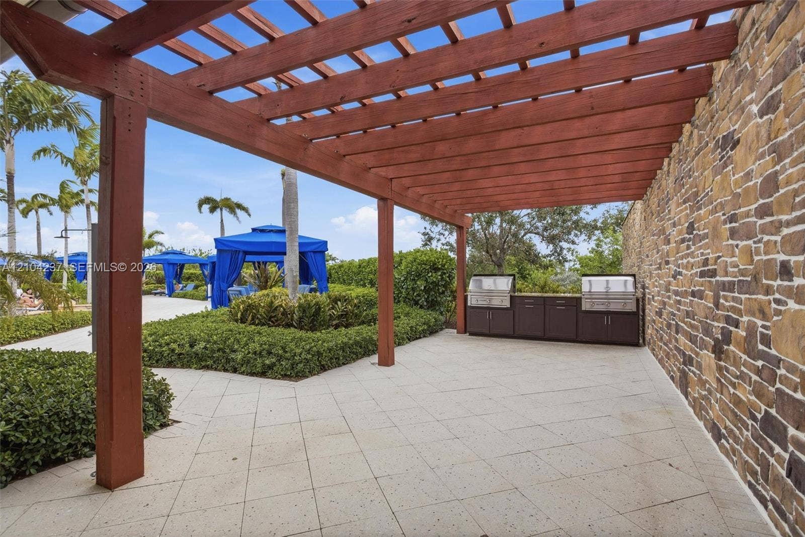 Outdoor living area featuring a wood-beam pergola, natural stone accent wall, built-in outdoor kitchen with stainless steel appliances, and paved patio flooring