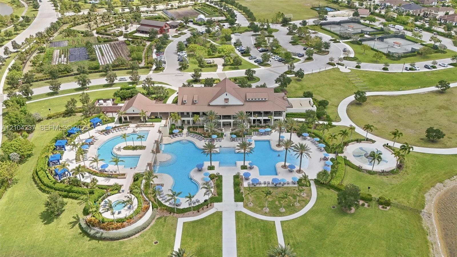 Aerial perspective of a multi-level clubhouse facility featuring an expansive resort-style swimming pool, integrated spa, paved sun deck, and surrounding manicured green space