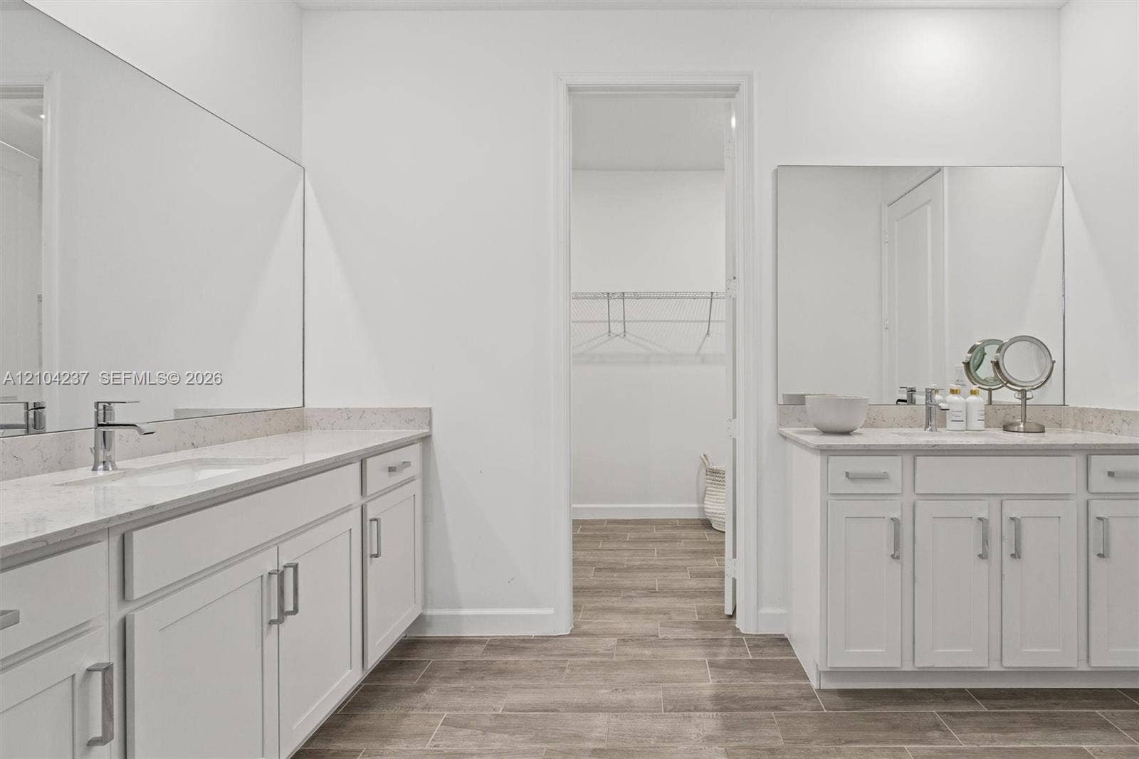 Dual vanity configuration featuring white Shaker-style cabinetry, light-toned composite countertops, large frameless mirrors, and wood-finish tile flooring