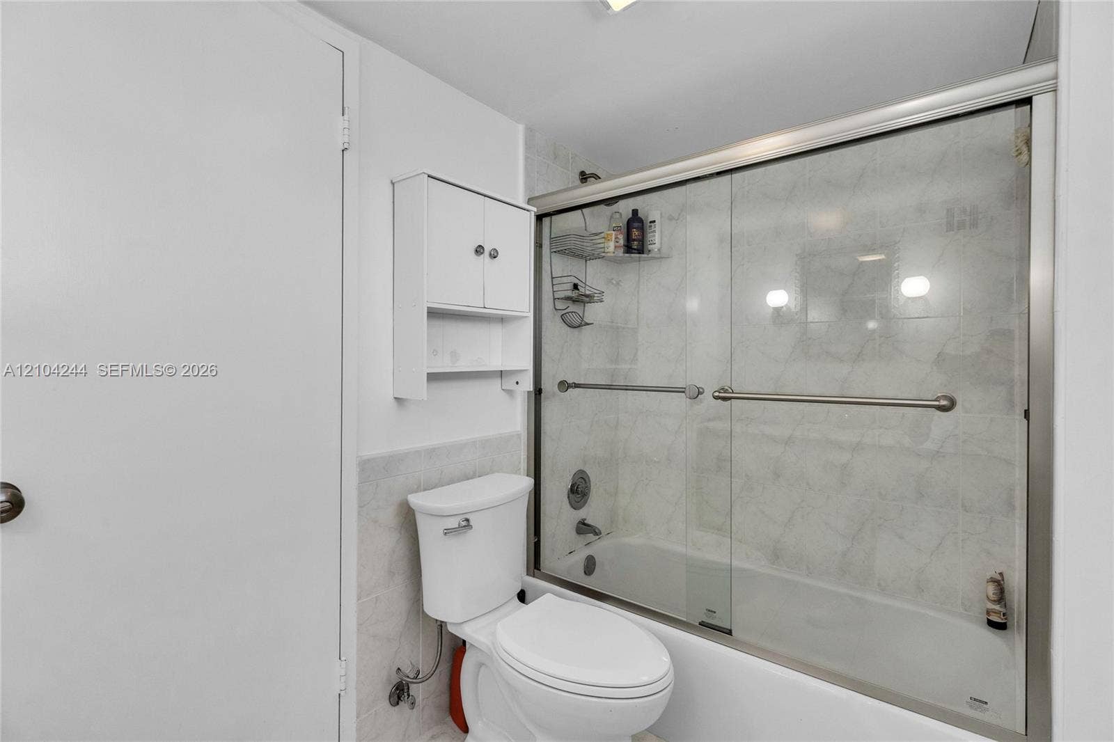 Bathroom featuring a white toilet, wall-mounted storage cabinet, and a bathtub with a glass sliding enclosure