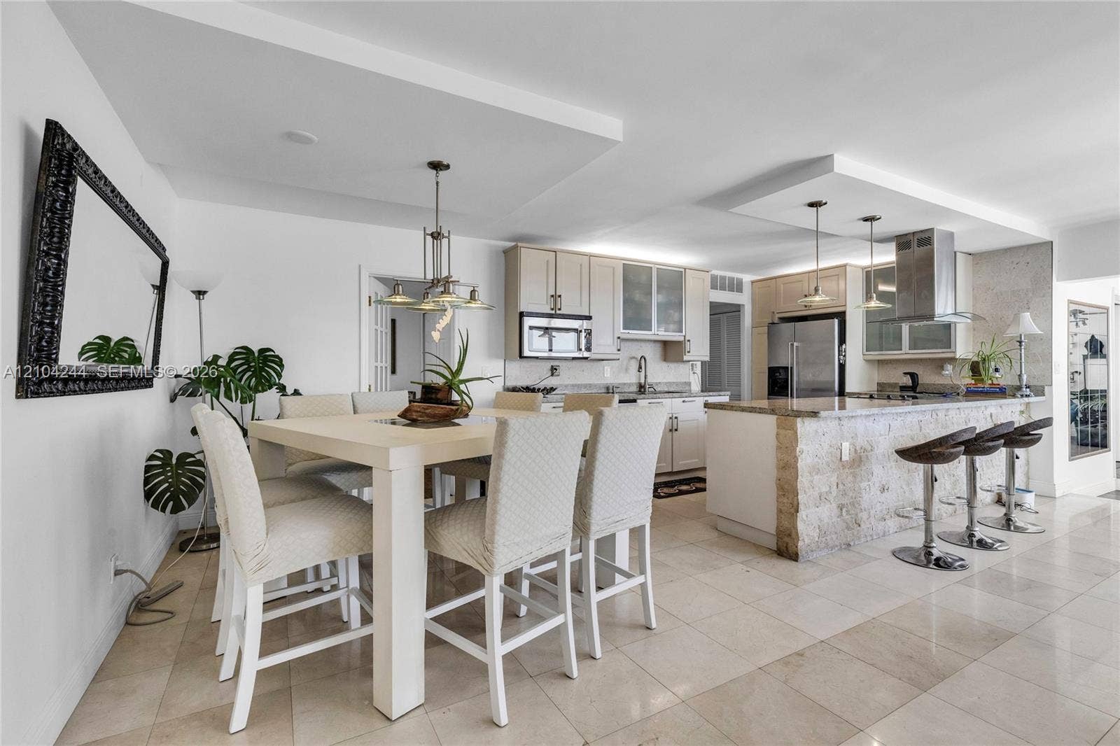 Open-concept kitchen featuring light wood cabinetry, stone-clad kitchen island, stainless steel appliances, tiered tray ceilings, and neutral tile flooring