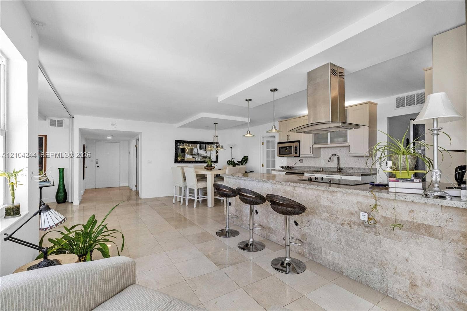 Open-concept kitchen featuring stone-clad breakfast bar, stainless steel chimney-style range hood, light-toned cabinetry, and tile flooring