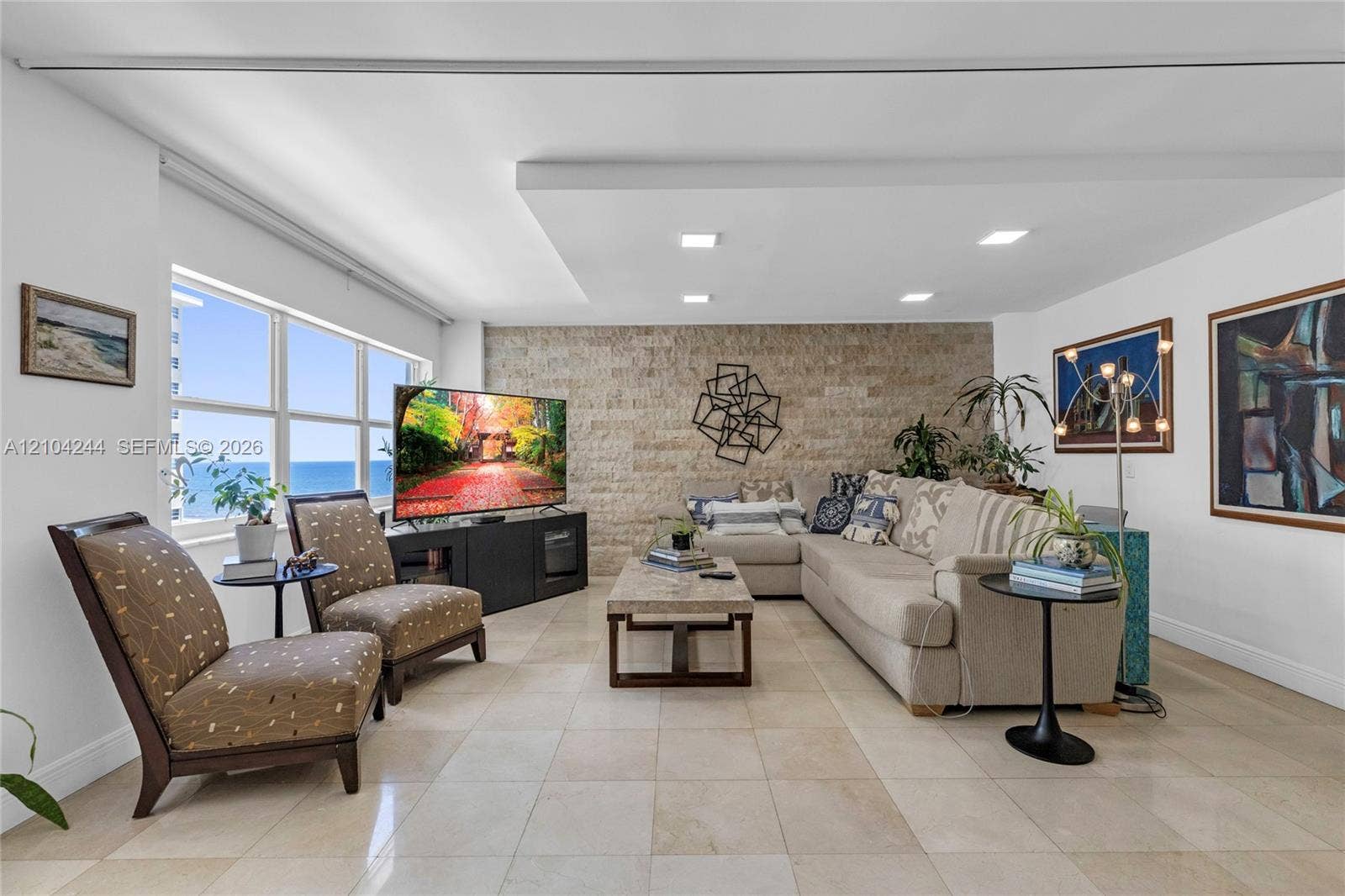 Living area featuring a textured stone accent wall, recessed ceiling lighting, neutral tile flooring, and large windows with water scenery