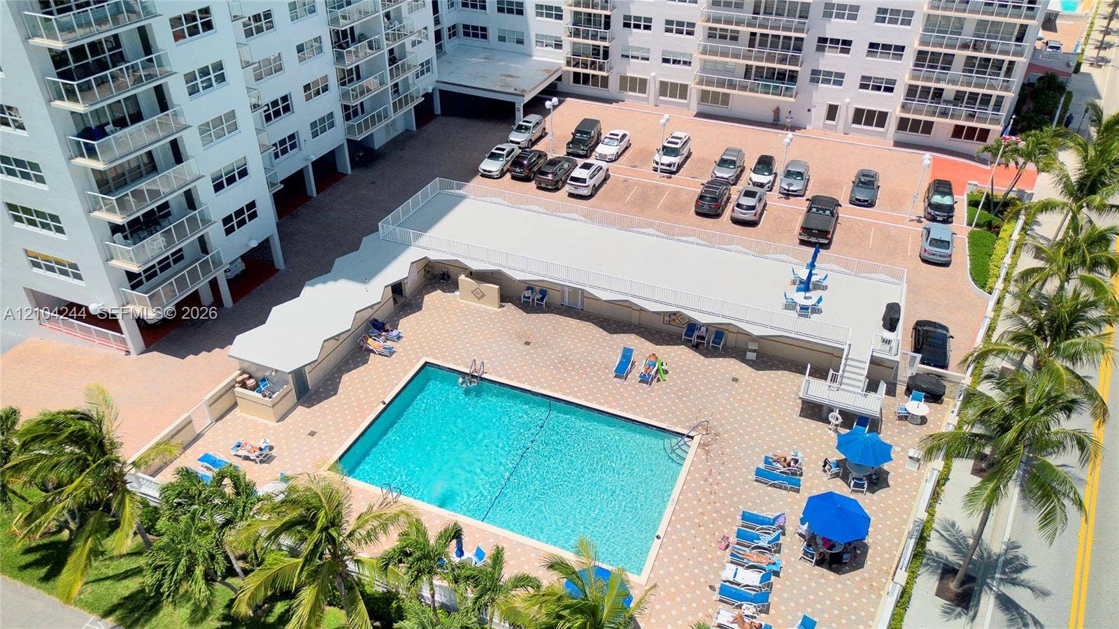 Aerial perspective of a multi-story residential complex featuring a paved parking area, a rectangular outdoor swimming pool, and concrete decking