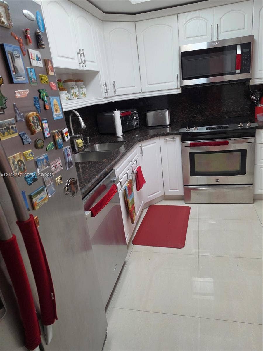 Kitchen featuring white cabinetry, dark stone-style countertops, stainless steel appliances, a double-basin undermount sink with a pull-down faucet, and polished tile flooring