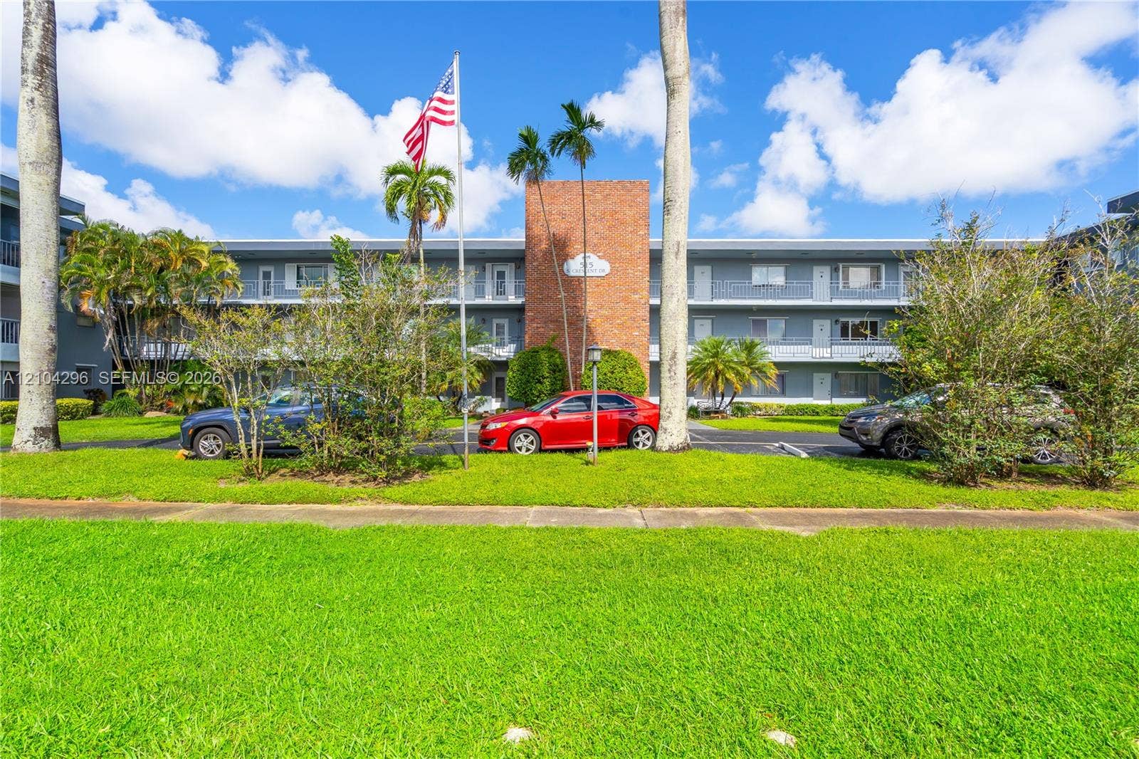 Mid-rise residential building exterior featuring a central red brick facade, grey siding, covered exterior walkways, and mature palm trees