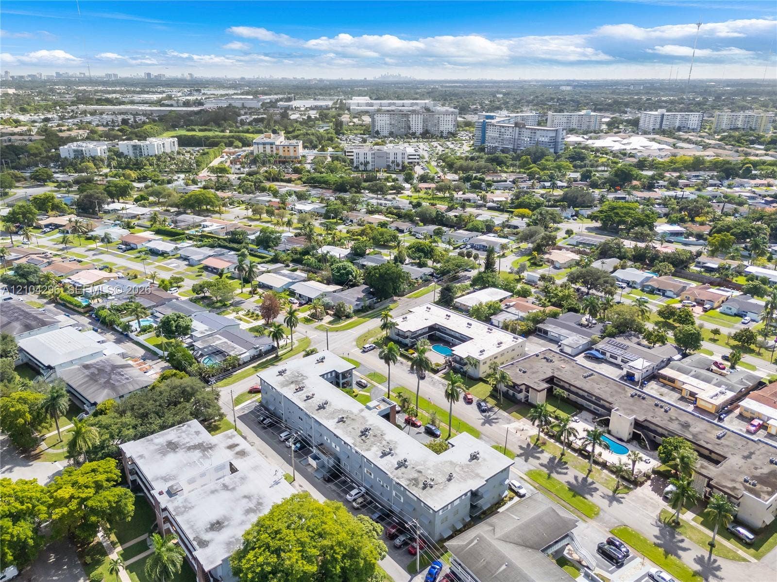 Aerial perspective of a multi-unit residential complex featuring flat roofs, central courtyards with swimming pools, and mature palm landscaping within a dense suburban grid