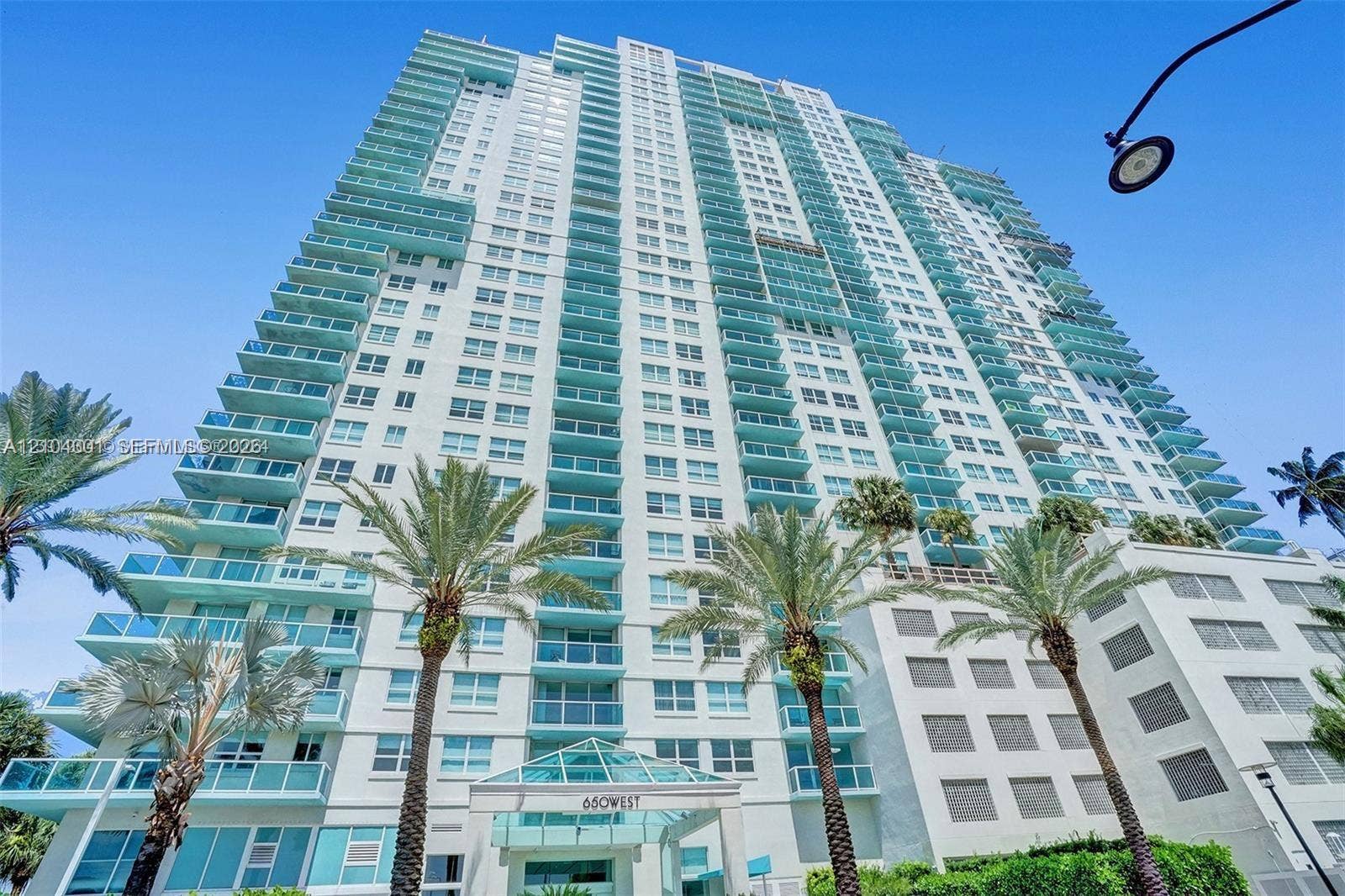 High-rise residential exterior featuring a light-toned facade, multiple levels of private glass-railed balconies, and a geometric glass-paneled entrance canopy, accented by mature palm trees