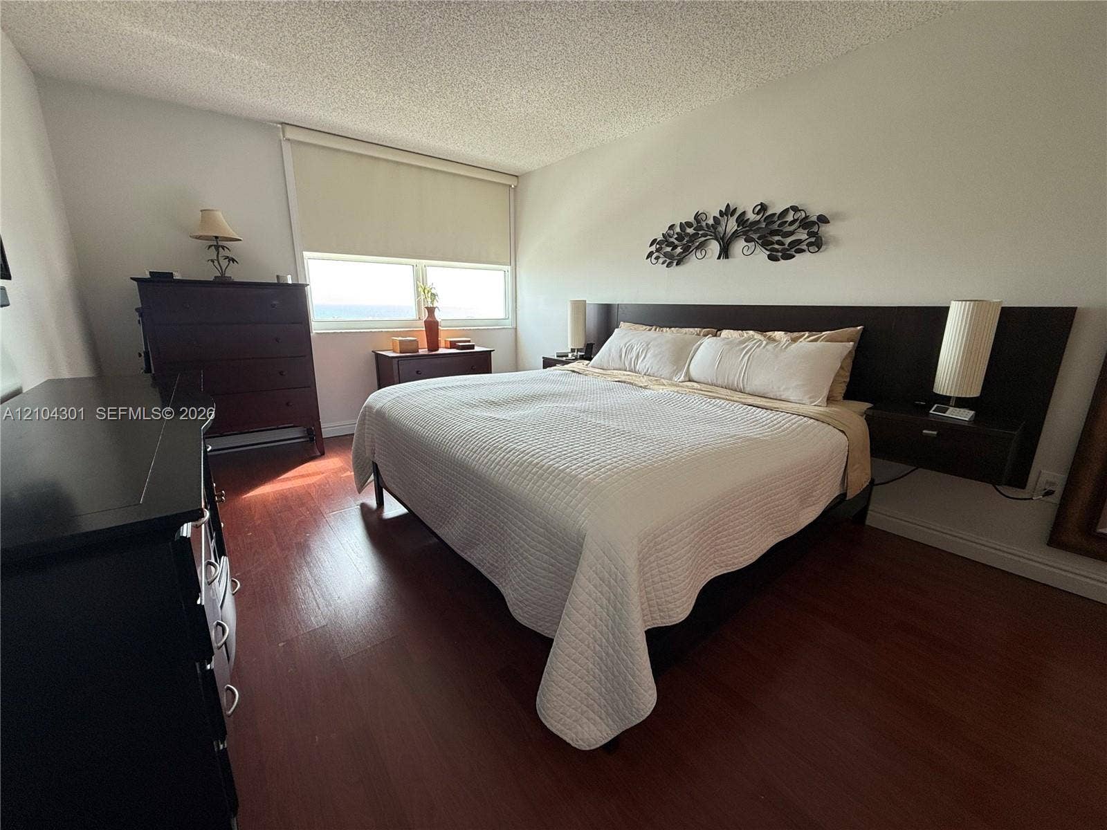 Bedroom featuring wood-finish flooring, a built-in dark wood headboard with integrated floating nightstands, and large windows with roller shades