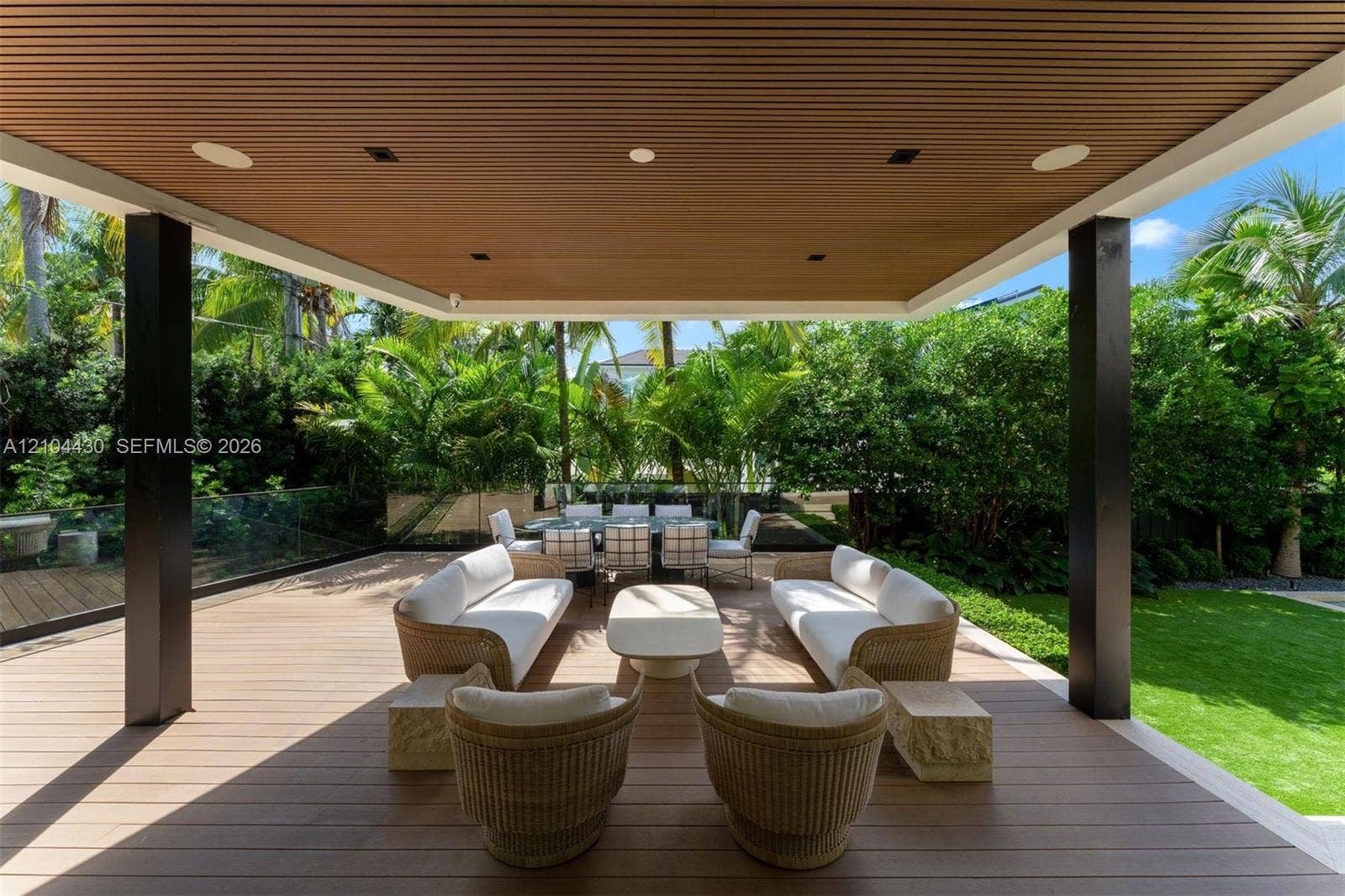 Covered patio with horizontal wood-slat ceiling, recessed lighting, composite decking, and glass panel railing overlooking a landscaped yard
