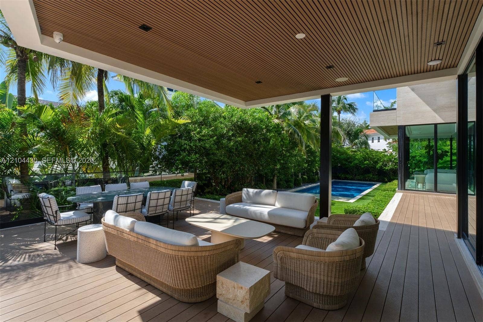 Covered outdoor patio with wood-slatted ceiling, composite decking, recessed lighting, glass railing, and in-ground pool