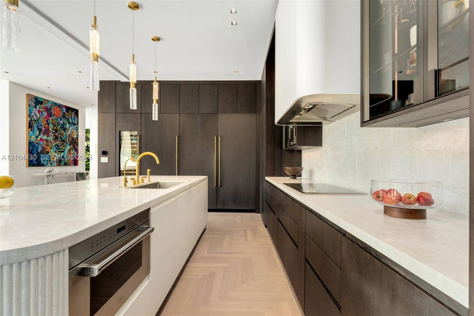 Gourmet kitchen featuring custom dark cabinetry, quartz countertops, a central island with a fluted pillar detail, herring-bone pattern wood-finish flooring, and integrated stainless steel appliances
