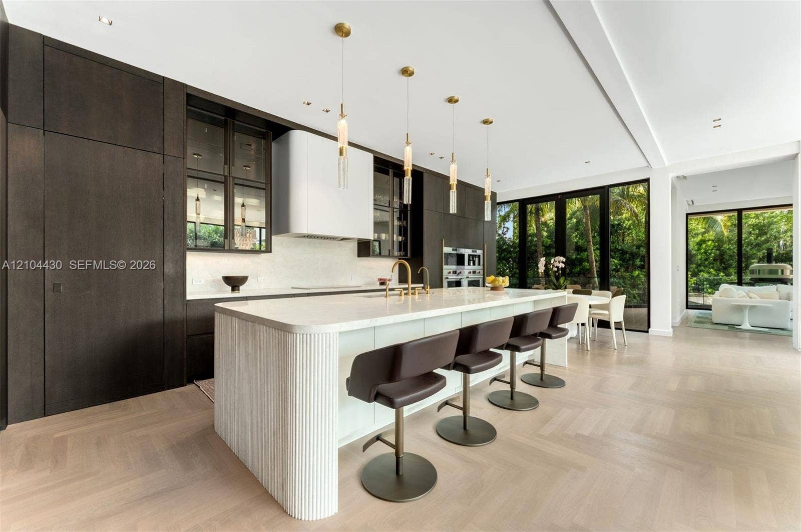 Gourmet kitchen featuring floor-to-ceiling dark wood cabinetry, a large central island with a fluted stone base, herringbone wood-finish flooring, built-in double ovens, and contemporary pendant lighting