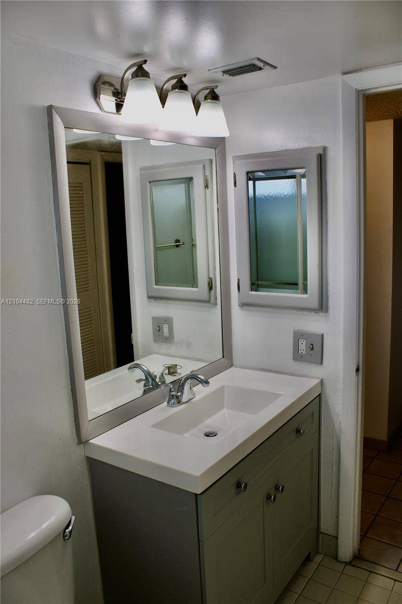 18000 NW 68th Avenue, #409A, Hialeah, Hialeah, FL 33015 Bathroom vanity featuring a solid-surface countertop with an integrated rectangular sink, three-light brushed-metal vanity fixture, framed wall mirror, and grey built-in storage cabinetry