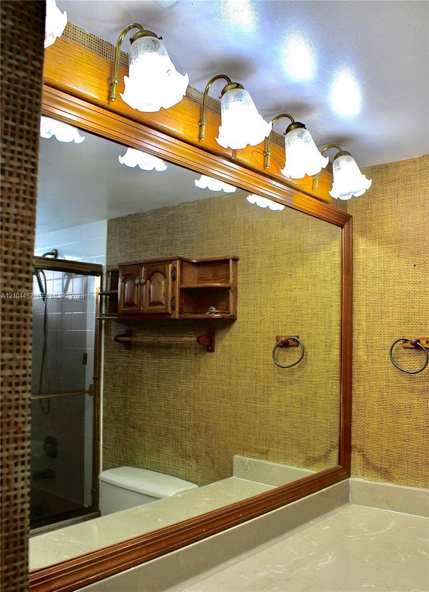 18000 NW 68th Avenue, #409A, Hialeah, Hialeah, FL 33015 Large, wood-framed wall mirror featuring a four-light fixture with glass shades, wall-mounted cabinetry, and a stone-finish vanity countertop