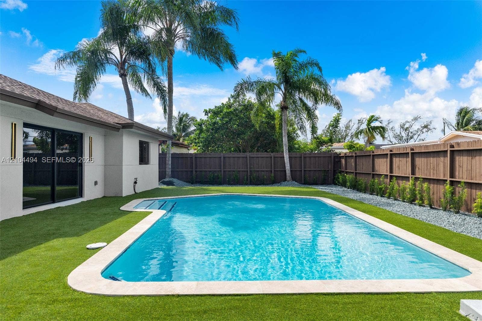 711 NW 8th Avenue, Dania Beach, FL 33004 Outdoor living area featuring a rectangular swimming pool with stone coping, synthetic turf, privacy wood fencing, and mature palm trees