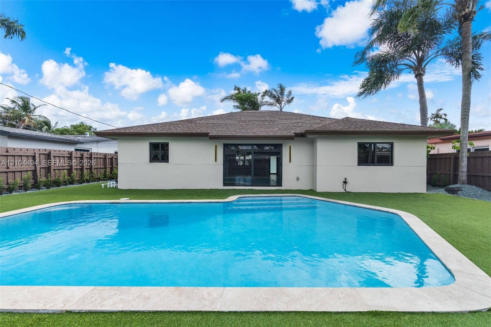 711 NW 8th Avenue, Dania Beach, FL 33004 Exterior rear elevation featuring a swimming pool with stone coping, synthetic turf lawn, privacy fencing, and sliding glass doors