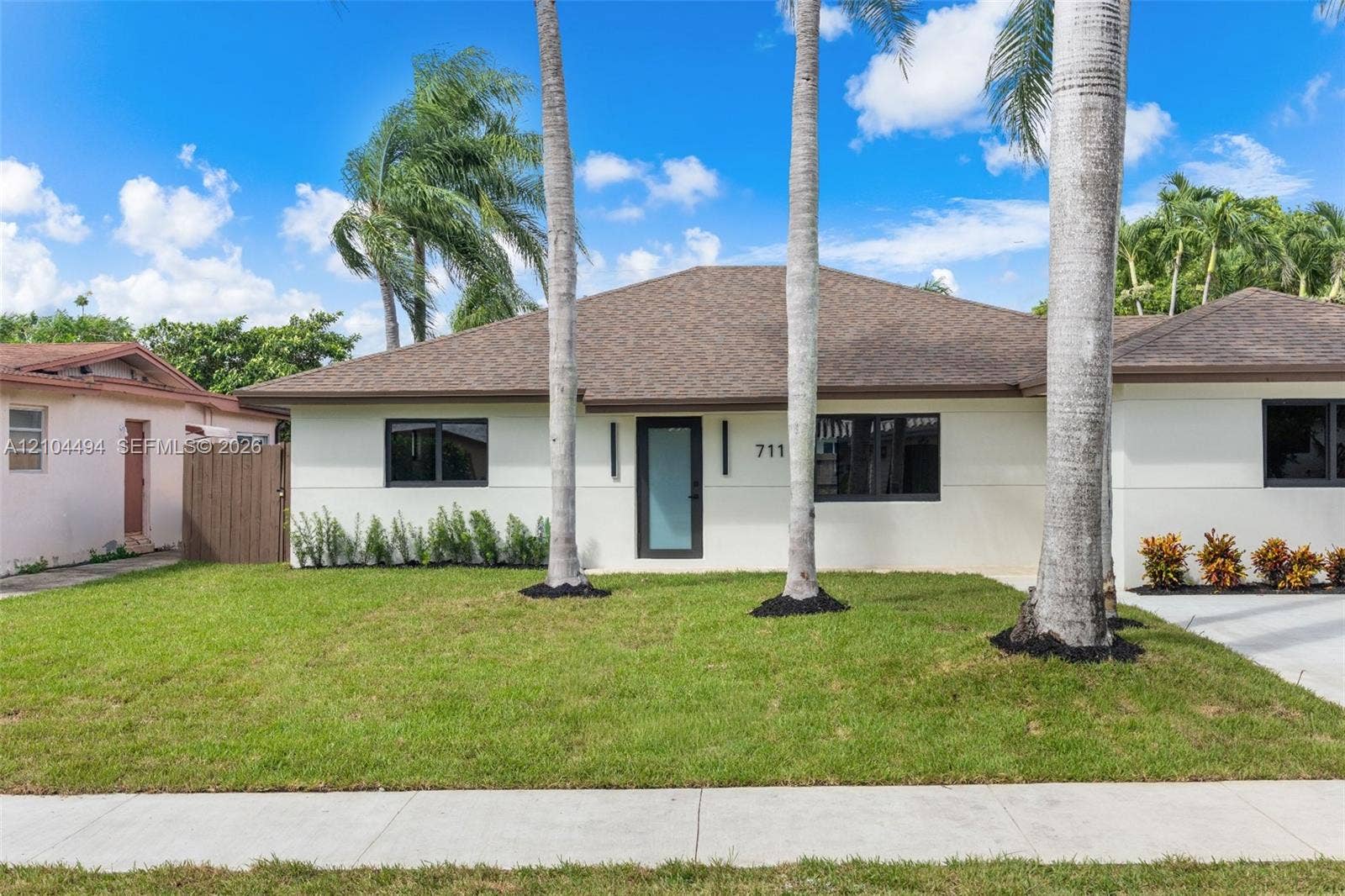 711 NW 8th Avenue, Dania Beach, FL 33004 Exterior featuring a light-colored stucco facade, a hipped roof with asphalt shingles, framed windows with dark trim, a glass-panel entry door, and manicured landscaping with mature palm trees