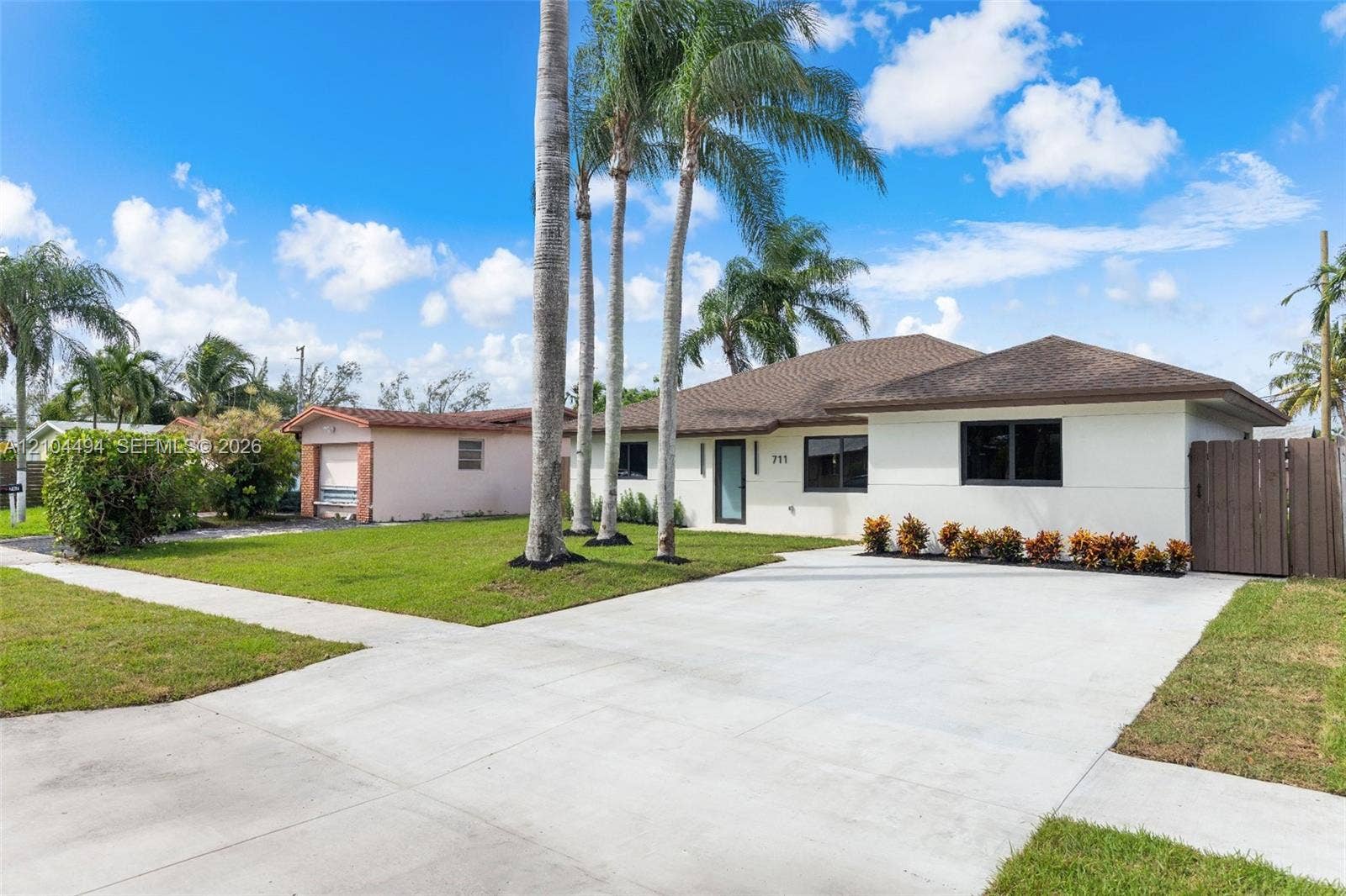 711 NW 8th Avenue, Dania Beach, FL 33004 Single-story residence featuring a neutral stucco exterior, a shingled hip roof, a concrete driveway, mature palm trees, and a wooden privacy gate