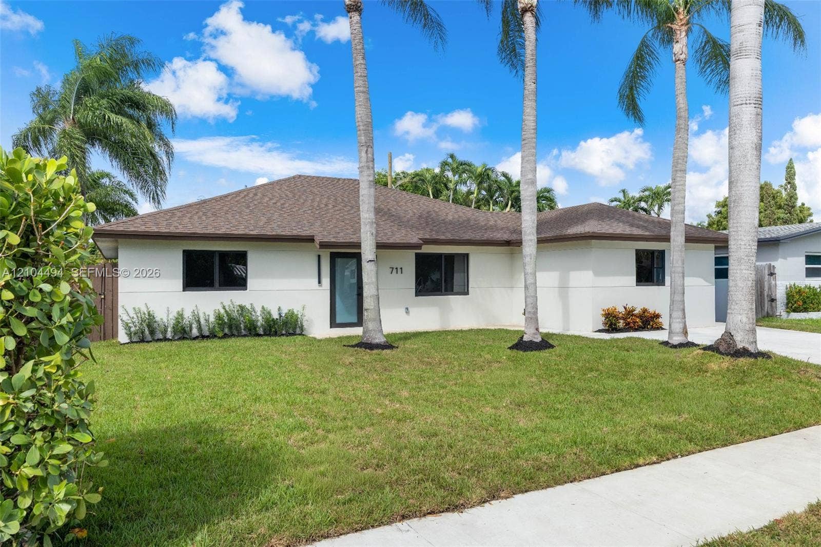 711 NW 8th Avenue, Dania Beach, FL 33004 Single-story exterior featuring a light-toned stucco faade, a hip roof, dark-framed windows, and mature palm trees