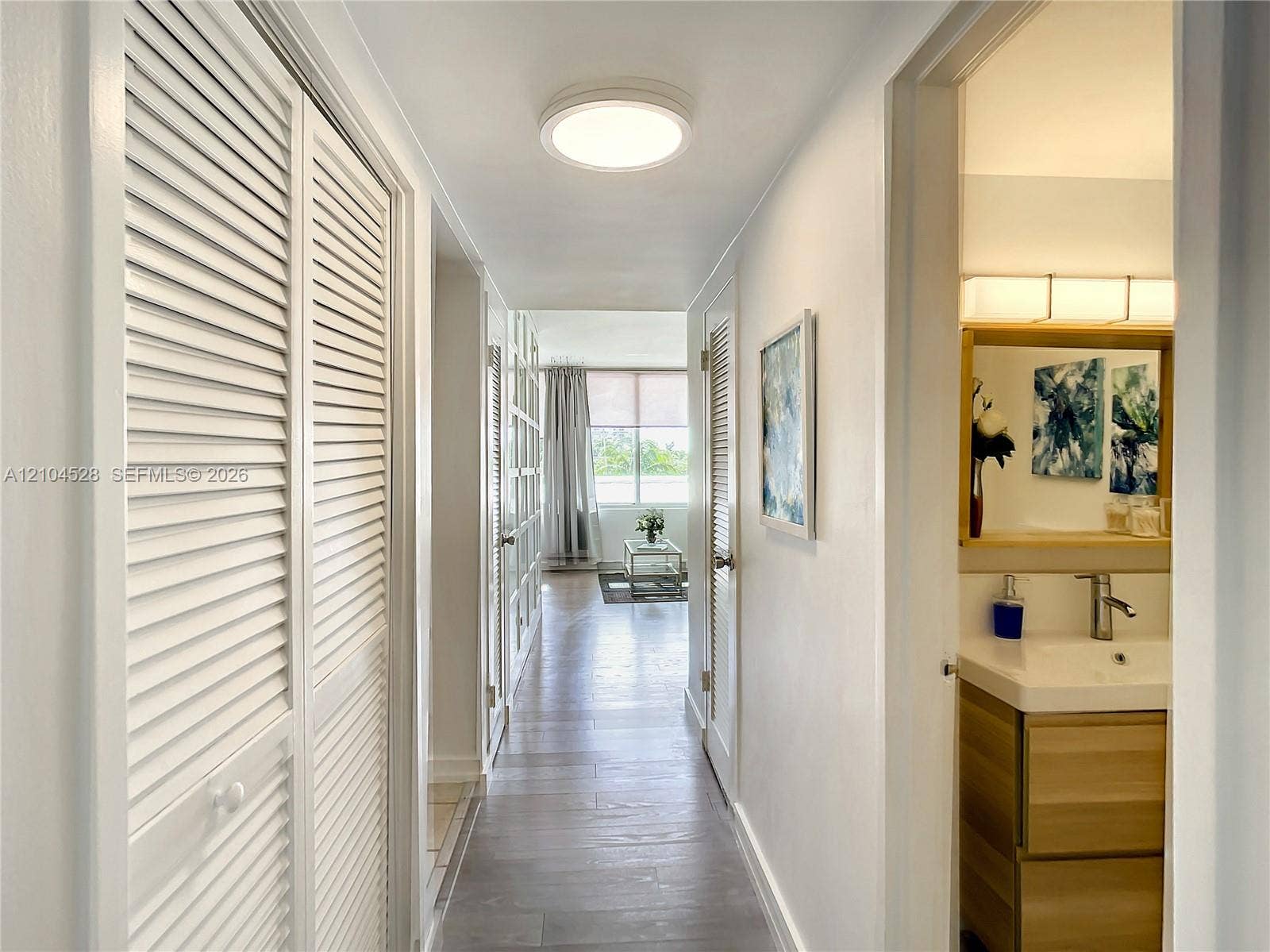 Hallway featuring wood-finish flooring, white louvered closet doors, and flush-mount lighting, with an entry into a bathroom showcasing a wood-finish vanity, integrated sink, and horizontal wall-mounted vanity light