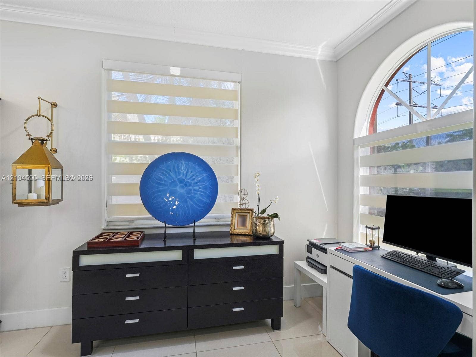 Arched window, tiered roller window shades, crown molding, gold-tone wall-mounted lantern light fixture, and neutral tile flooring