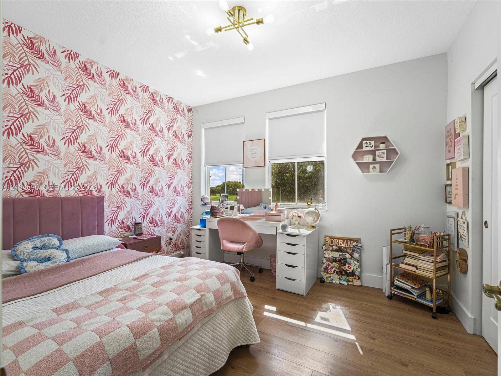 Bedroom featuring wood-finish flooring, dual windows with roller shades, a gold-tone multi-arm chandelier, and a decorative patterned accent wall
